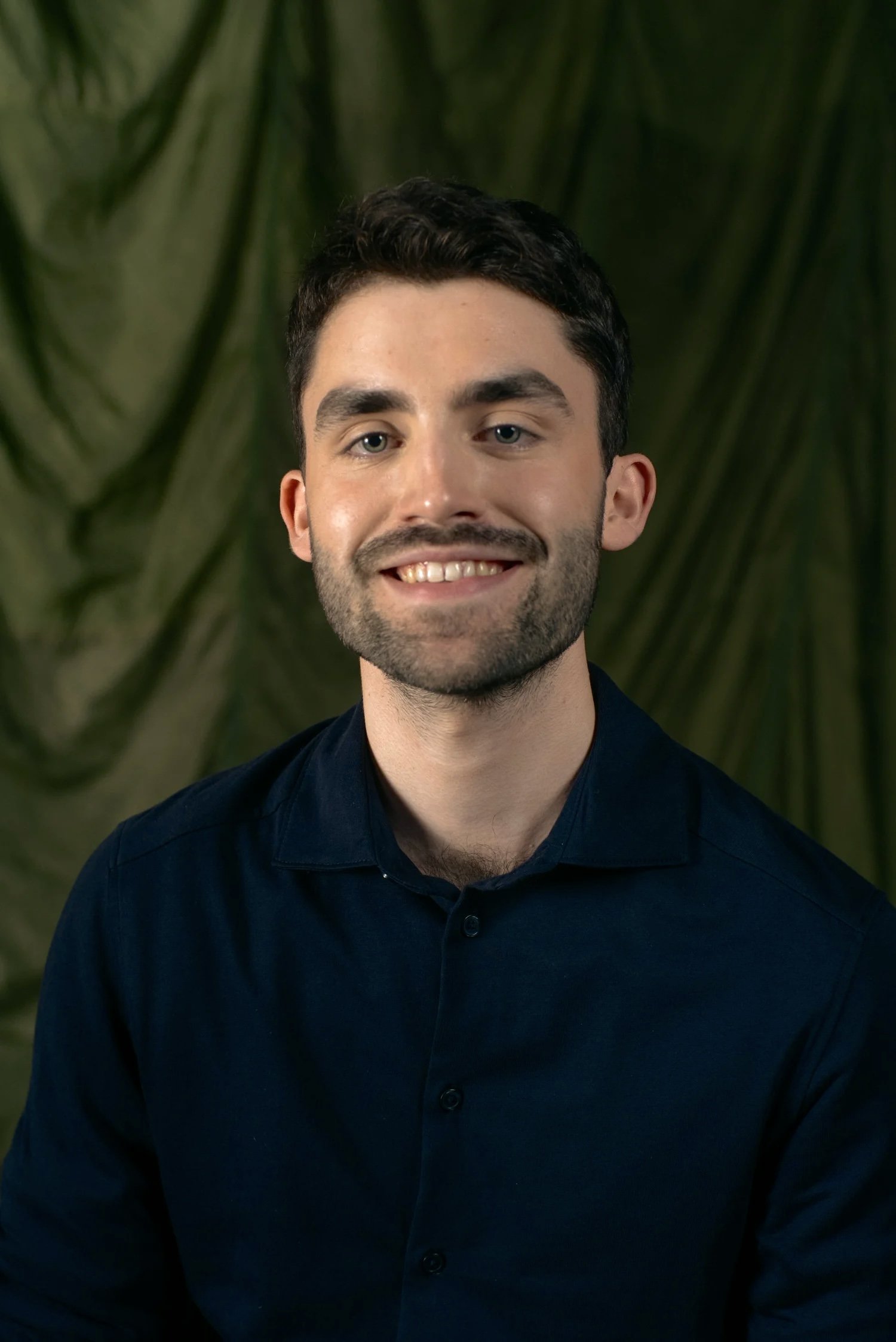 Close-up portrait of a young man with dark hair, light blue eyes, and a short beard, smiling, wearing a dark blue shirt, with green leafy plants blurred in the background.
