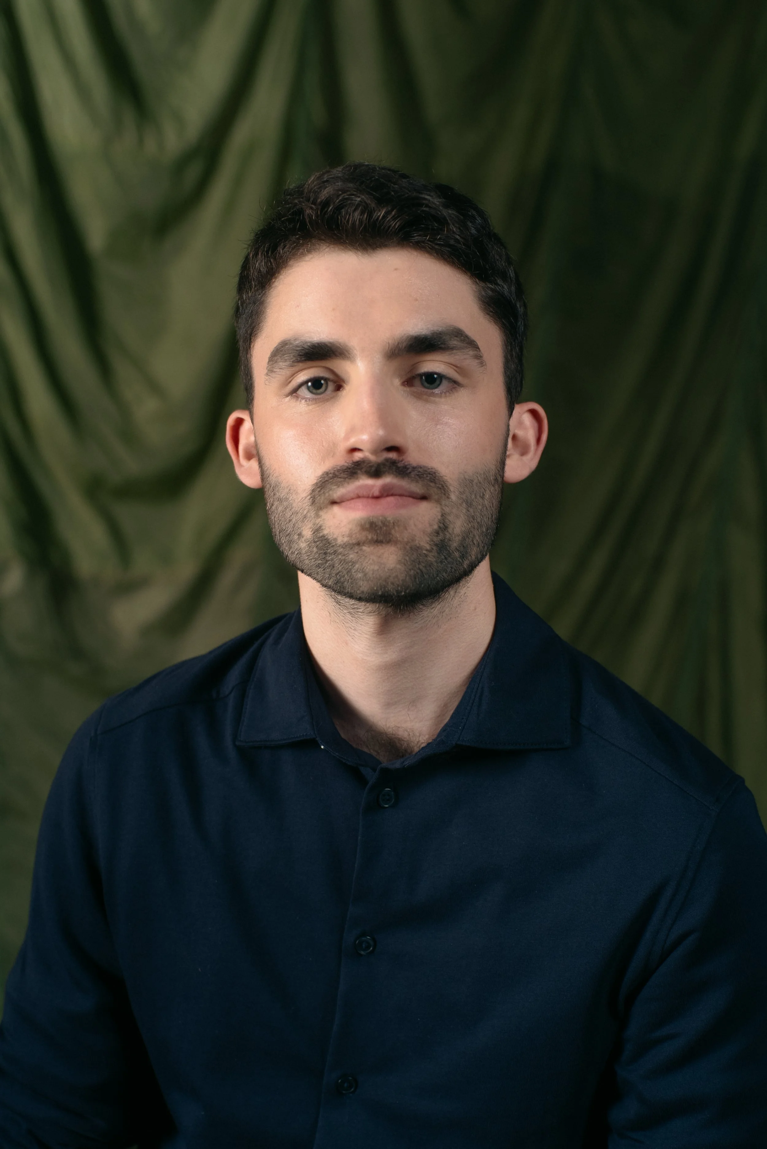 A young man with dark hair, beard, and blue eyes wearing a dark navy button-up shirt, posed against a green leafy background.