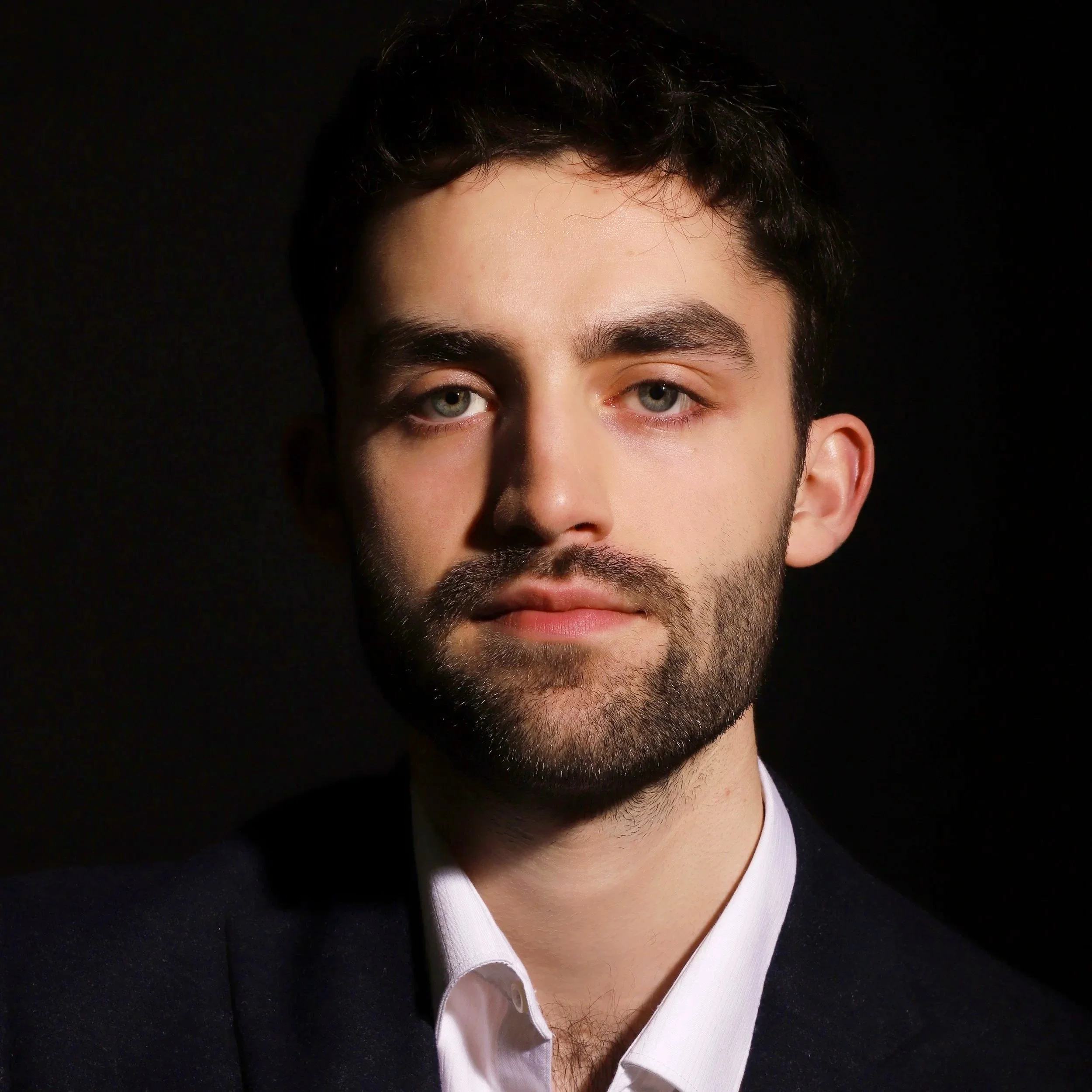 Close-up of a man's face, showing his eyes, eyebrows, and part of his nose, with dark hair and a beard, wearing a white shirt and a dark jacket against a black background.