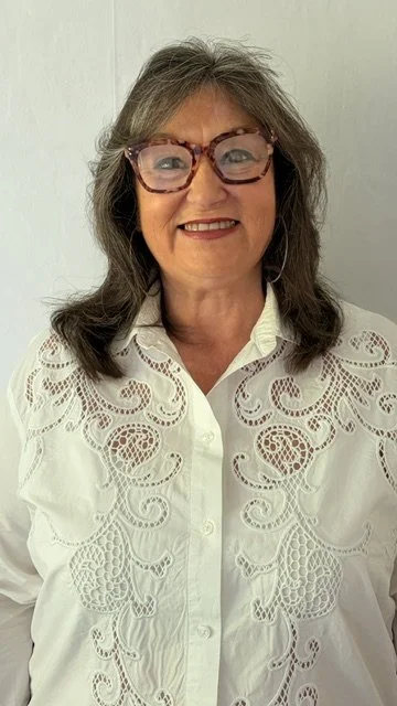 A smiling woman with gray hair wearing on-trend tortoise-shaped glasses and a white embroidered blouse against a plain light background.