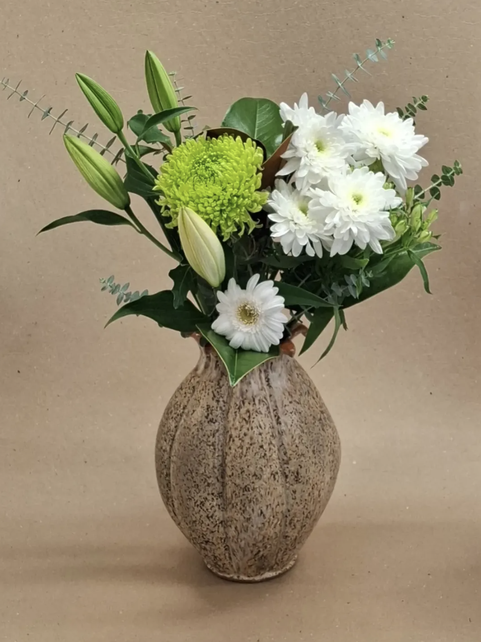 A beige textured ceramic vase holding a bouquet of white chrysanthemums, green lilies, and various greenery against a plain background.