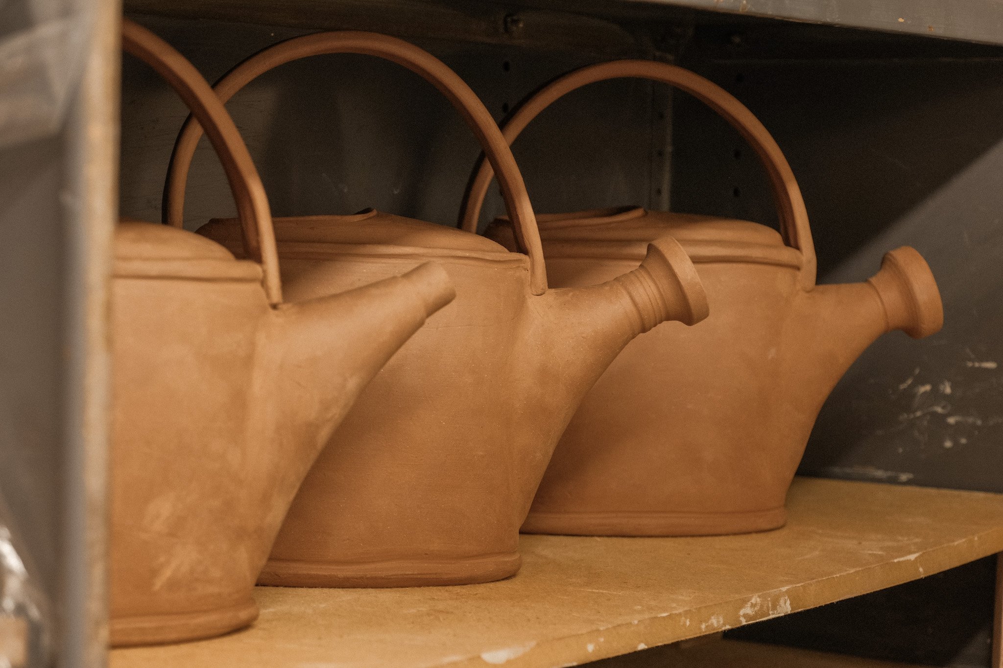 Three unglazed ceramic watering cans with arched handles on a wooden shelf.