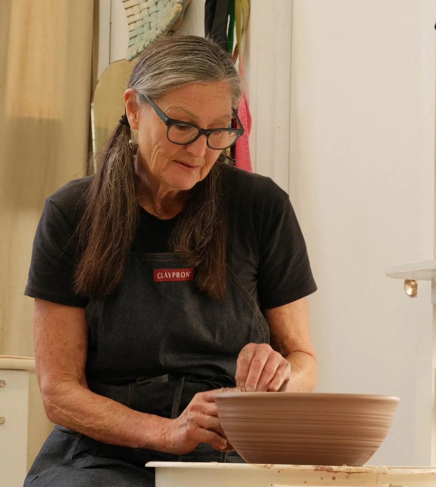 An elderly woman with gray hair tied in pigtails wearing glasses and a black t-shirt with a red 'Claypron' patch, working on a pottery wheel making a ceramic bowl.