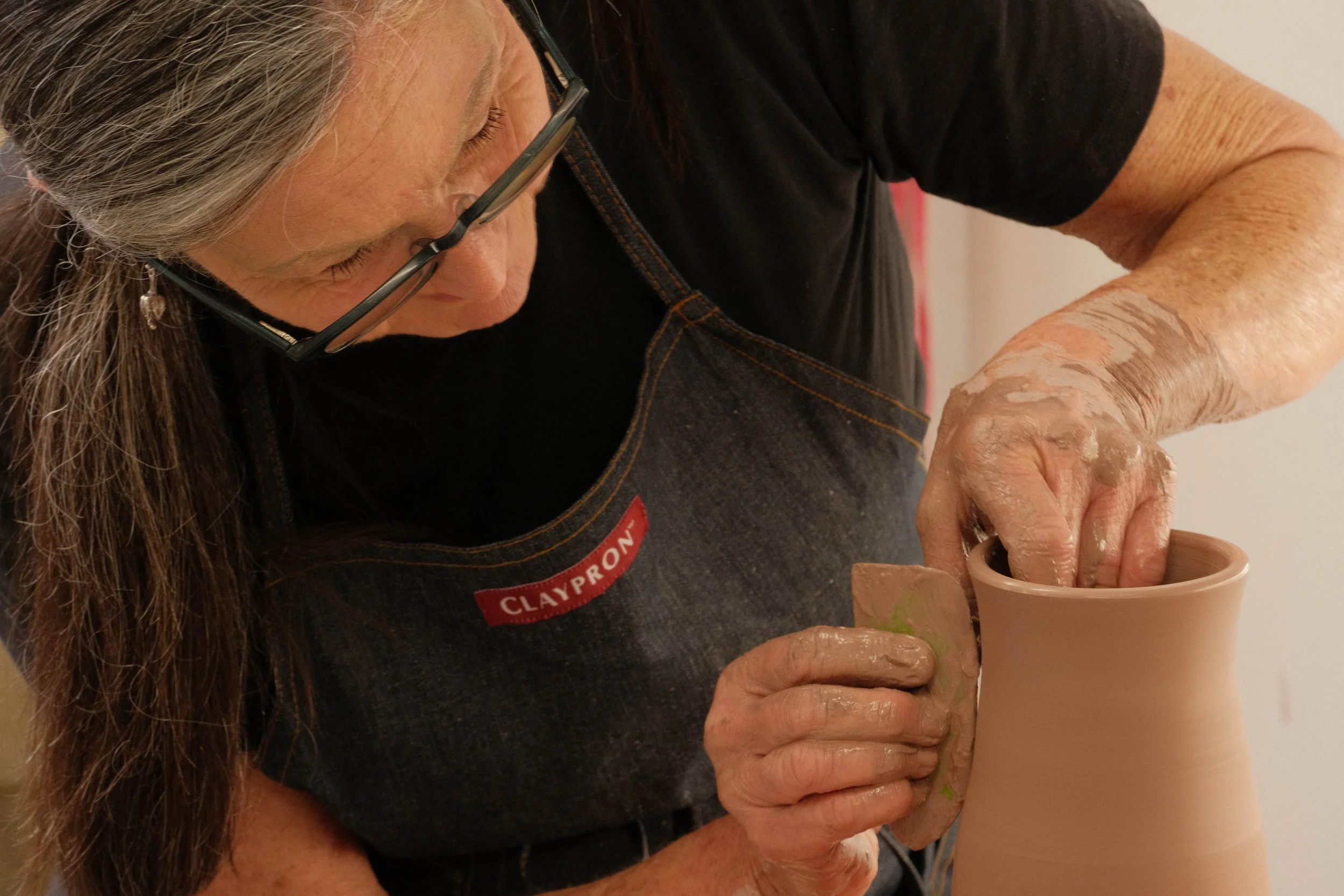 An elderly woman with glasses crafting pottery on a pottery wheel, wearing a black shirt and a brown apron with a red name tag that reads "Claypro".