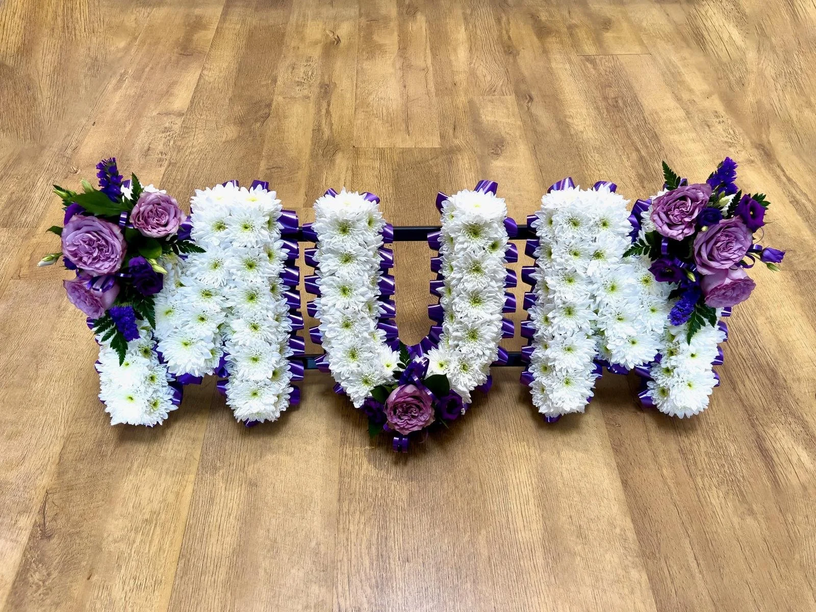 
"MUM" Lavender Rose and Rich Purple Tribute | A beautiful and regal funeral tribute spelling  out "MUM". The arrangement features letters densely based with crisp white chrysanthemums, sharply outlined by a striking, deep purple pleated ribbon borde