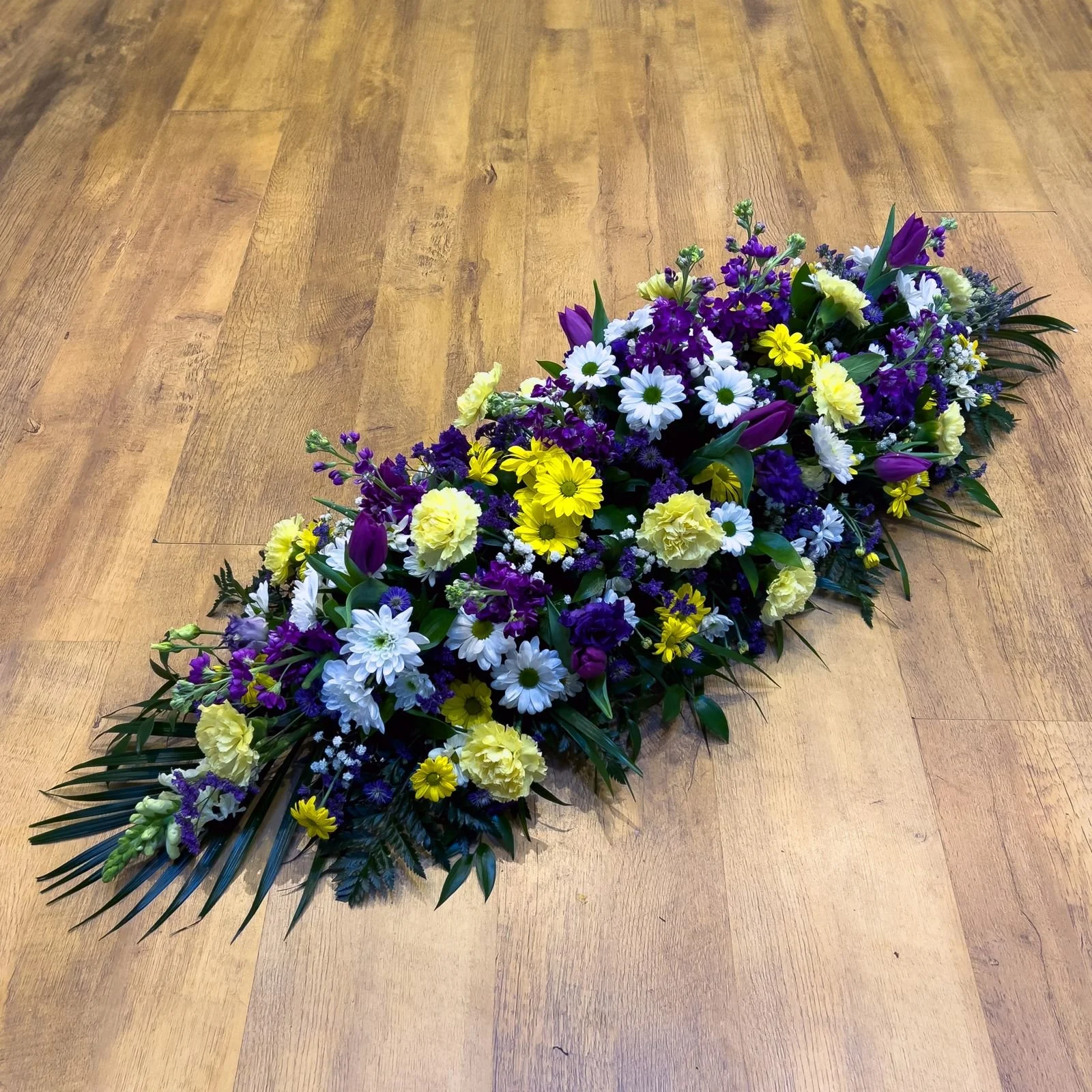 Cheerful Yellow and Purple Spray | A bright and uplifting tribute that pairs contrasting tones beautifully. This spray features cheerful yellow carnations, vibrant yellow chrysanthemum, and bold purple tulips, interspersed with deep purple stocks and