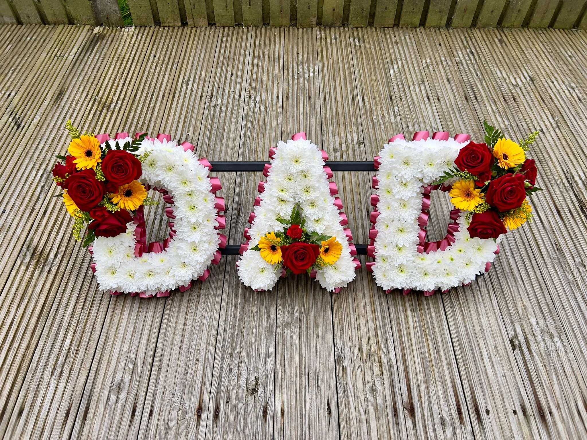 "DAD" Vibrant Red and Yellow Gerbera Tribute | Built upon a clean white chrysanthemum base, the letters are sharply defined by a pleated burgandy ribbon border. High-contrast floral sprays featuring fiery red roses and cheerful yellow gerbera daisies