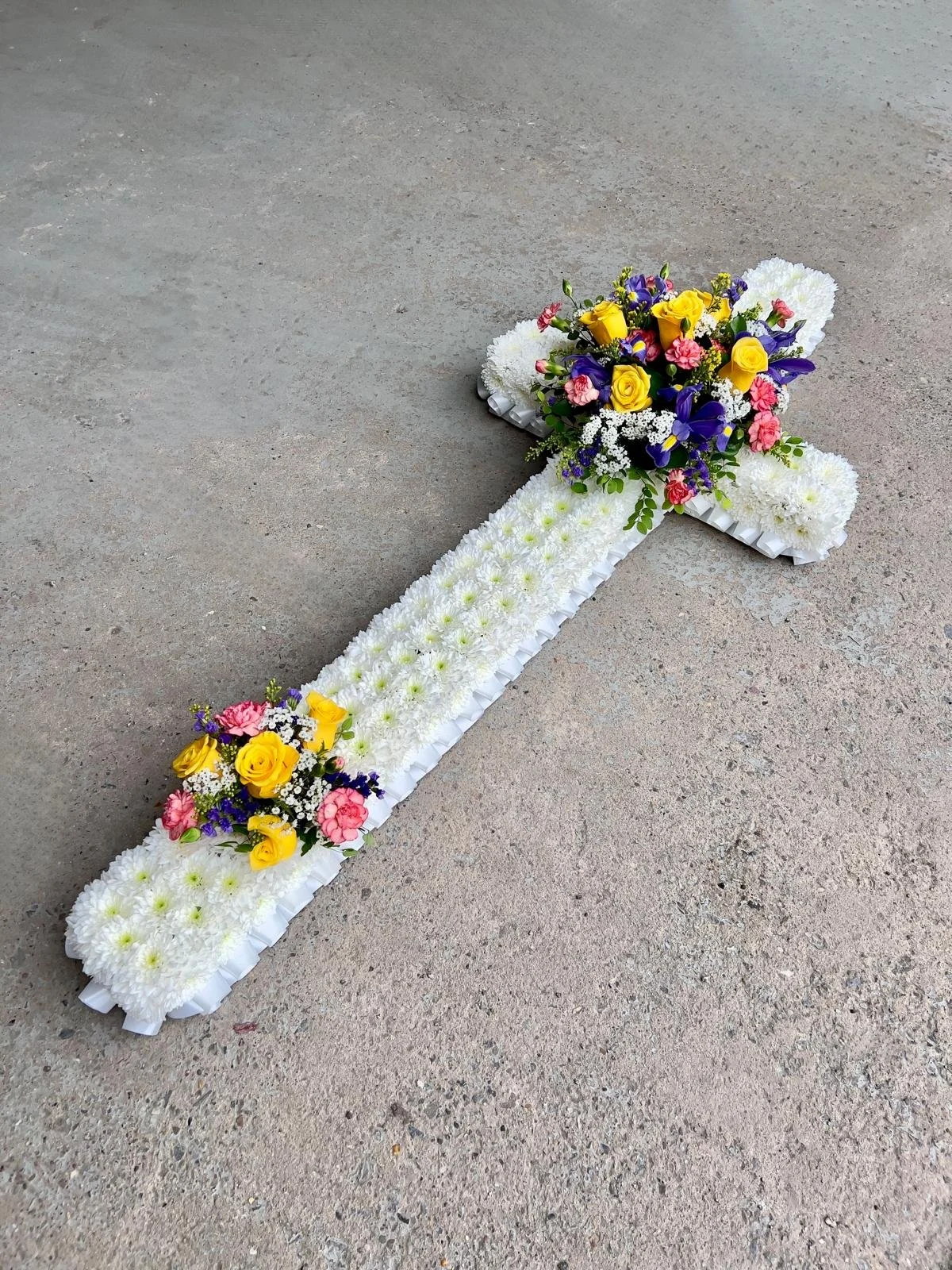 Vibrant Yellow and Purple Floral Cross Tribute | A beautifully structured, traditional funeral cross arrangement. The cross is intricately based with tight white chrysanthemums and finished with a clean, white pleated ribbon border. It is strikingly 