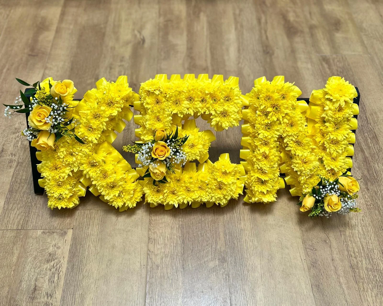 "KEN" Monochromatic Canary Yellow Tribute | A bright and unforgettable tribute spelling "KEN". Perfect for celebrating a life full of joy, this arrangement is entirely massed in vibrant natural canary-yellow chrysanthemums and finished with a matchin