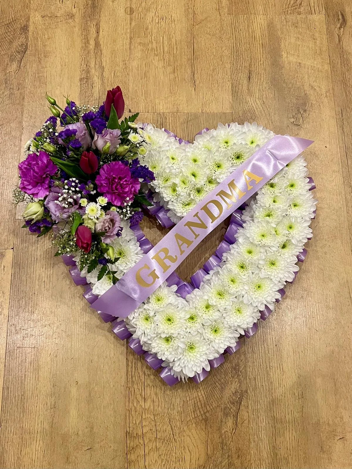 Lavender Open Heart Tribute | A tender, open-heart floral tribute dedicated to a beloved grandmother. The heart shape is neatly constructed from white chrysanthemums and edged with a lavender pleated ribbon. A soft purple banner reading "GRANDMA" str