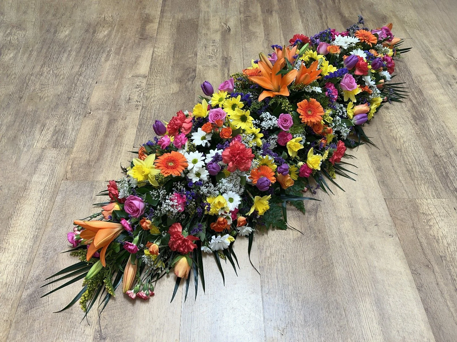 Vibrant Kaleidoscope Spray | A magnificent, high-density explosion of color that beautifully honors a vibrant life. This spray features striking orange lilies, bright red carnations, orange gerberas, sunny yellow daisies, pink roses, and purple tulip