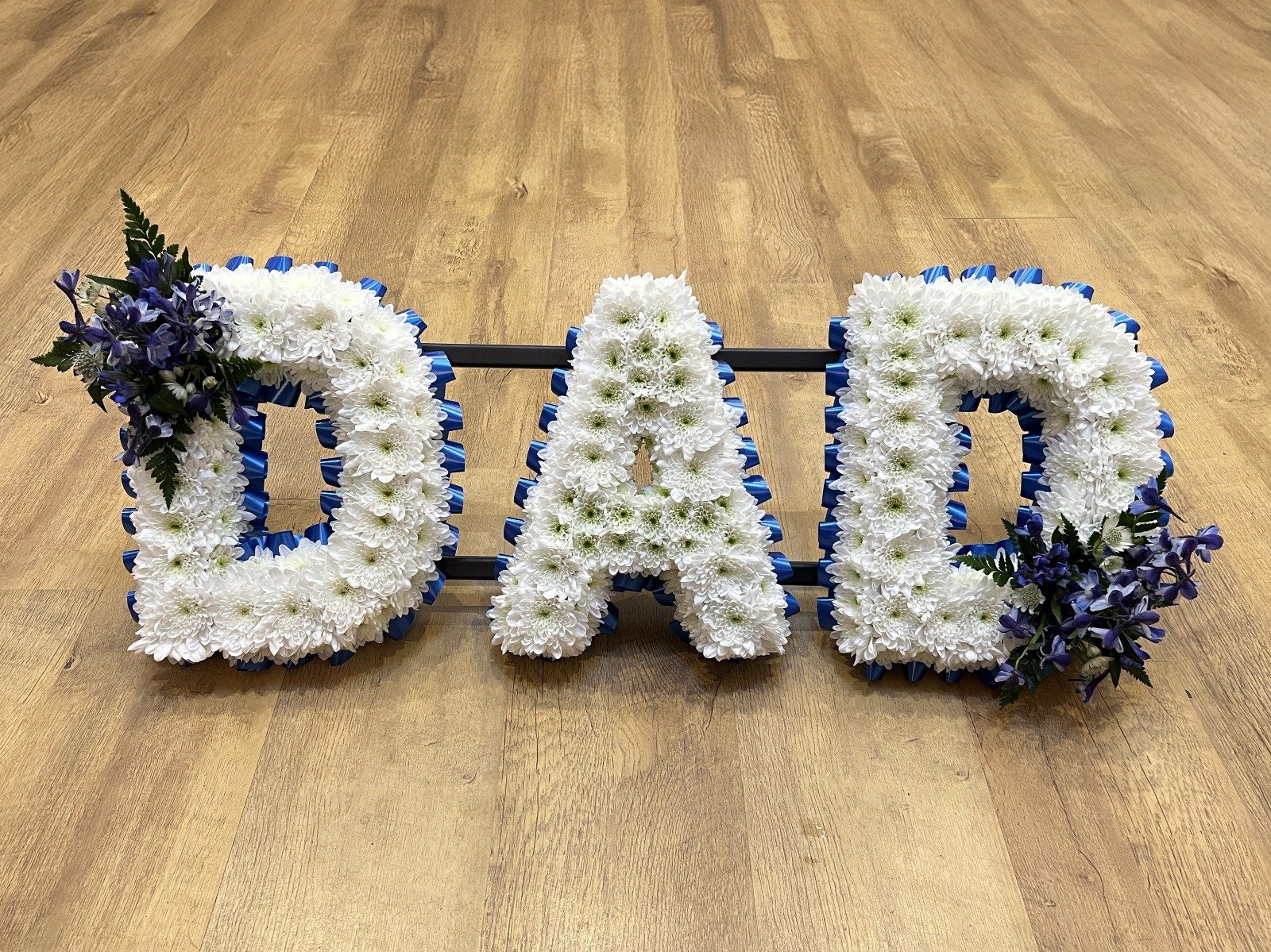 "DAD" Deep Blue and White Tribute | A stately and dignified tribute spelling "DAD". The stark white chrysanthemum letters are vividly framed by a royal blue pleated ribbon border, giving the arrangement an impactful silhouette. Rich clusters of midni