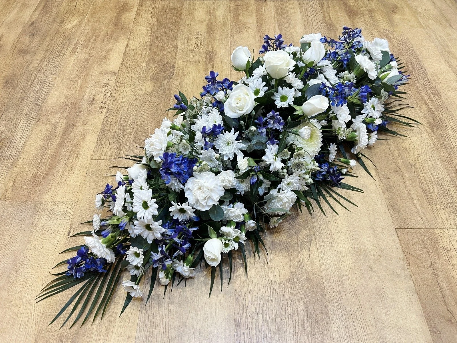 Classic Navy Blue and White Spray | A sophisticated, high-contrast tribute featuring elegant white roses, white carnations, and white daisies paired with rich, deep blue delphiniums and statice. The bold blue pops beautifully against the clean white 
