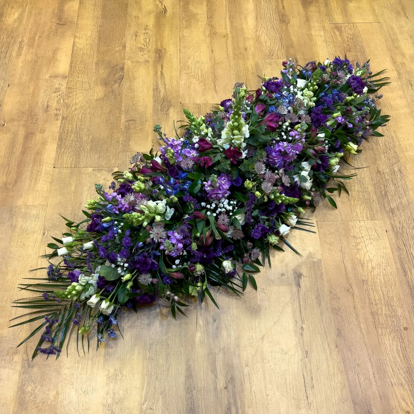 Deep Purple and Lilac Meadow Spray | A lush, meadow-style arrangement focused heavily on a rich spectrum of purples. It brings together deep violet lisianthus, lavender stocks, clusters of purple veronica, and white snapdragons, subtly accented by de