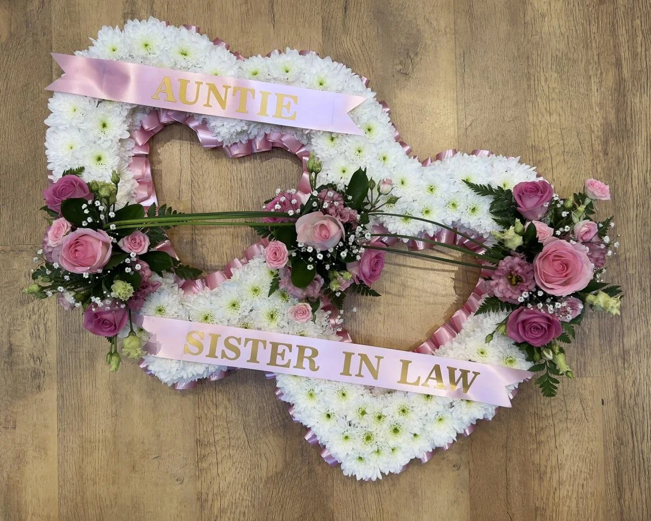 Double Open Heart | A touching interlocking double-heart tribute expressing dual devotion. Each open heart is massed in soft white chrysanthemums, lined with a delicate pastel-pink pleated ribbon. Elegant floral sprays of romantic pink roses, pink li