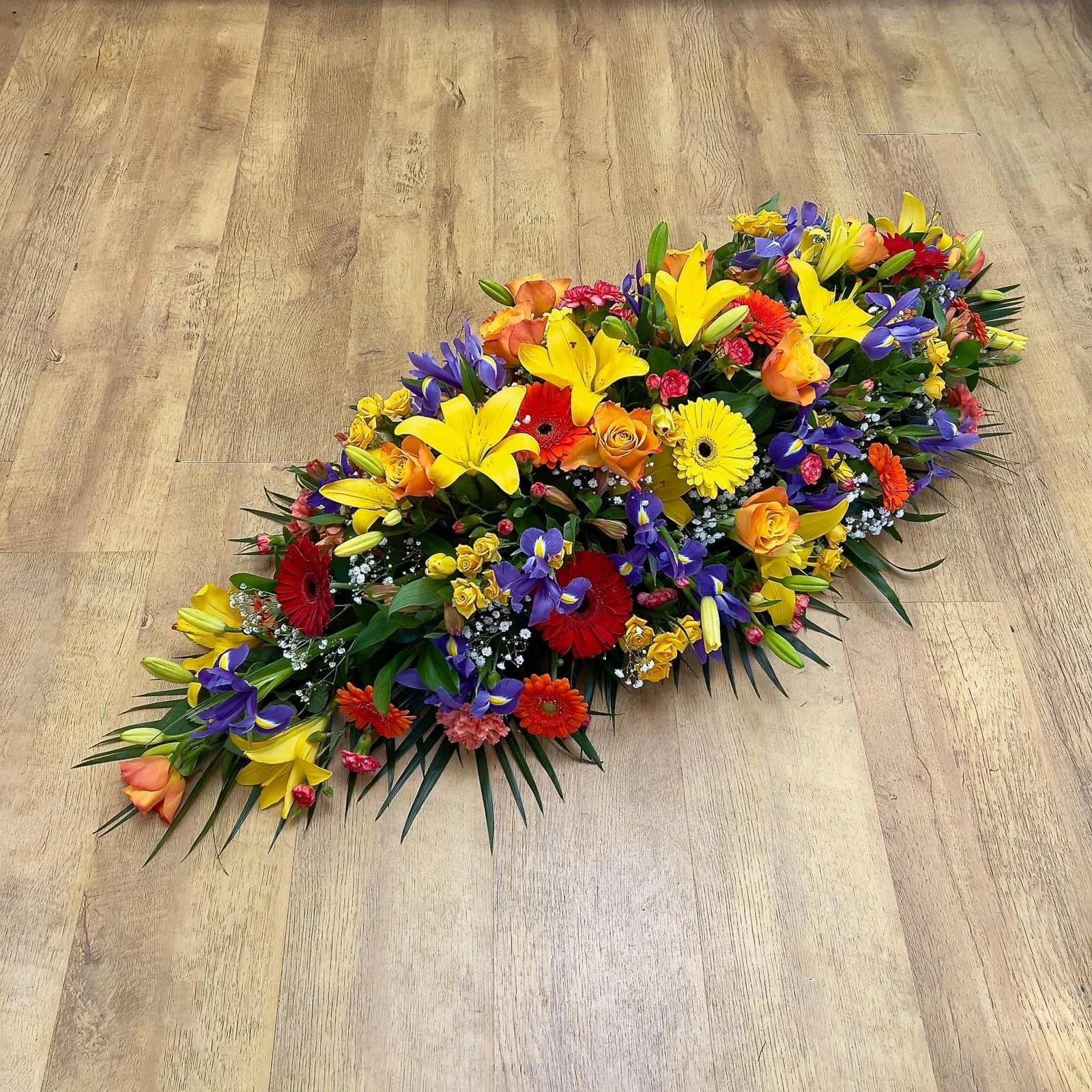 Vibrant Mix with Iris Spray | Celebratory, this vibrant arrangement is filled withyellow lilies, bright orange roses, and fiery red gerbera daisies. Striking purple irises provide a beautiful contrast, making this a popular colorful tribute.