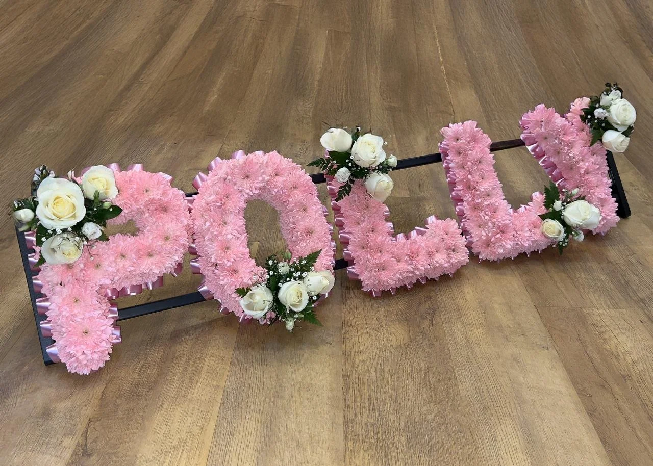 "POLLY" Soft Pink Rose Tribute | Each letter is tightly based with sprayed soft pink chrysanthemums and neatly edged with a coordinating pink pleated ribbon border. The tribute is elegantly broken up by alternating sprays of crisp white roses and del