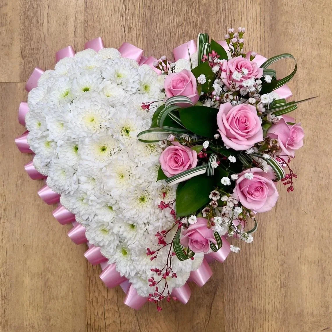 Heart-shaped floral arrangement with white chrysanthemums and pink roses, decorated with pink ribbon and greenery on a wooden surface.