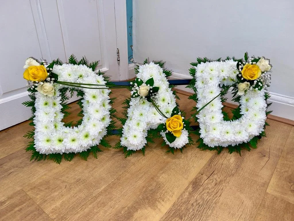 "DAD" Golden Yellow and Cream Rose Tribute | A peaceful and bright tribute spelling out "DAD". Each letter features a plush base of pristine white chrysanthemums set against a soft frame of natural green foliage. The arrangement is delicately connect