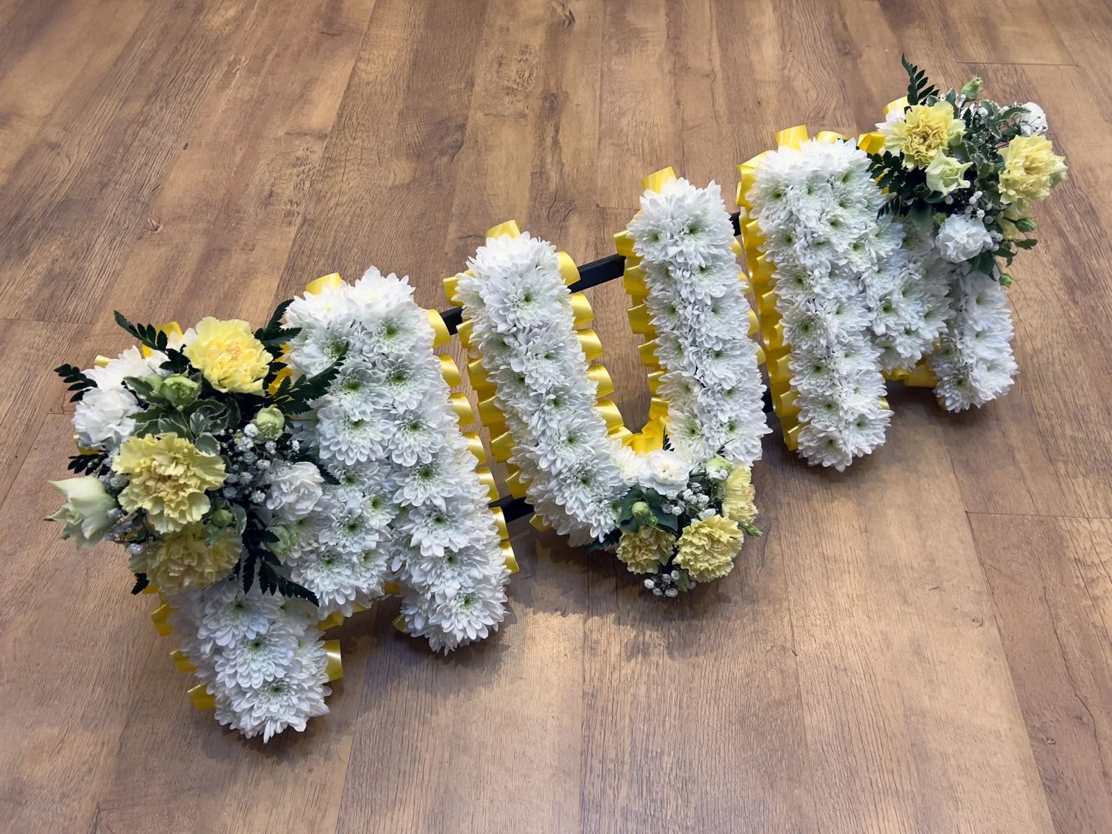 "MUM" Lemon and Yellow Carnation Tribute | A cheerful, multi-layered tribute spelling "MUM". The letters feature a solid base of white chrysanthemums accented by a vibrant yellow pleated ribbon border. Lush focal clusters of lemon-yellow carnations, 