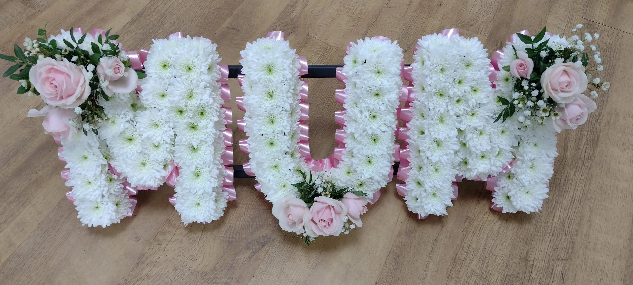 Soft Pink Rose & White Tribute | A tender and classic tribute spelling "MUM". The pristine white chrysanthemum letters are surrounded by a soft pastel-pink pleated ribbon border. Delicate, romantic sprays of pale pink roses, spray roses, and airy bab
