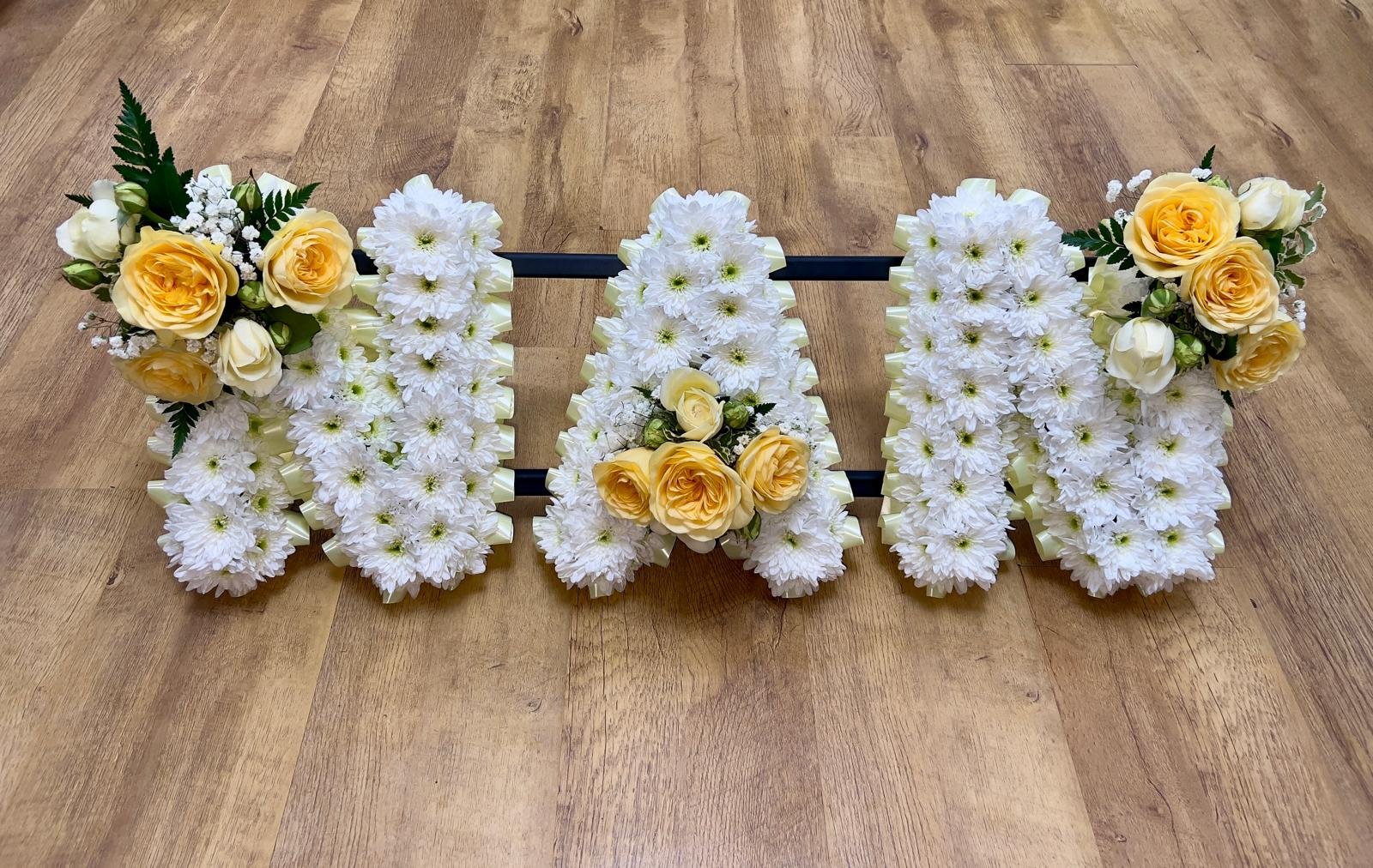 "NAN" Yellow Rose Tribute | A soft, vintage-inspired tribute spelling "NAN". Each letter is solidly massed with white chrysanthemums and finished with a delicate cream-colored pleated ribbon border. Warm, romantic sprays of yellow roses and white spr
