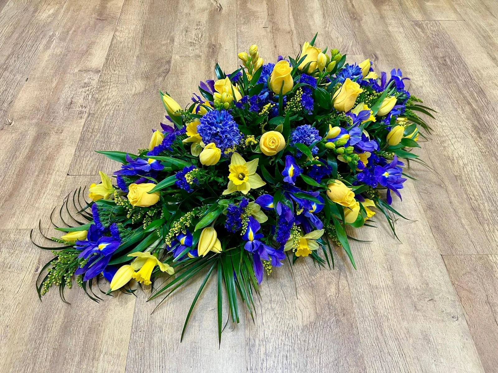 Springtime Yellow and Sapphire Blue Spray | A vibrant and deeply comforting springtime tribute that beautifully balances contrast. This elegant arrangement showcases a sunny selection of bright yellow roses, classic daffodils, and slender yellow tuli
