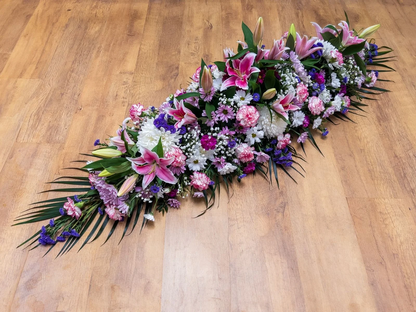 Vibrant Pink, Purple & White Mixed Spray | A detailed and texturally rich display containing striking pink lilies, soft pink carnations, and multi-petaled white chrysanthemums. Splashes of purple statice and delicate lavender daisies weave through th