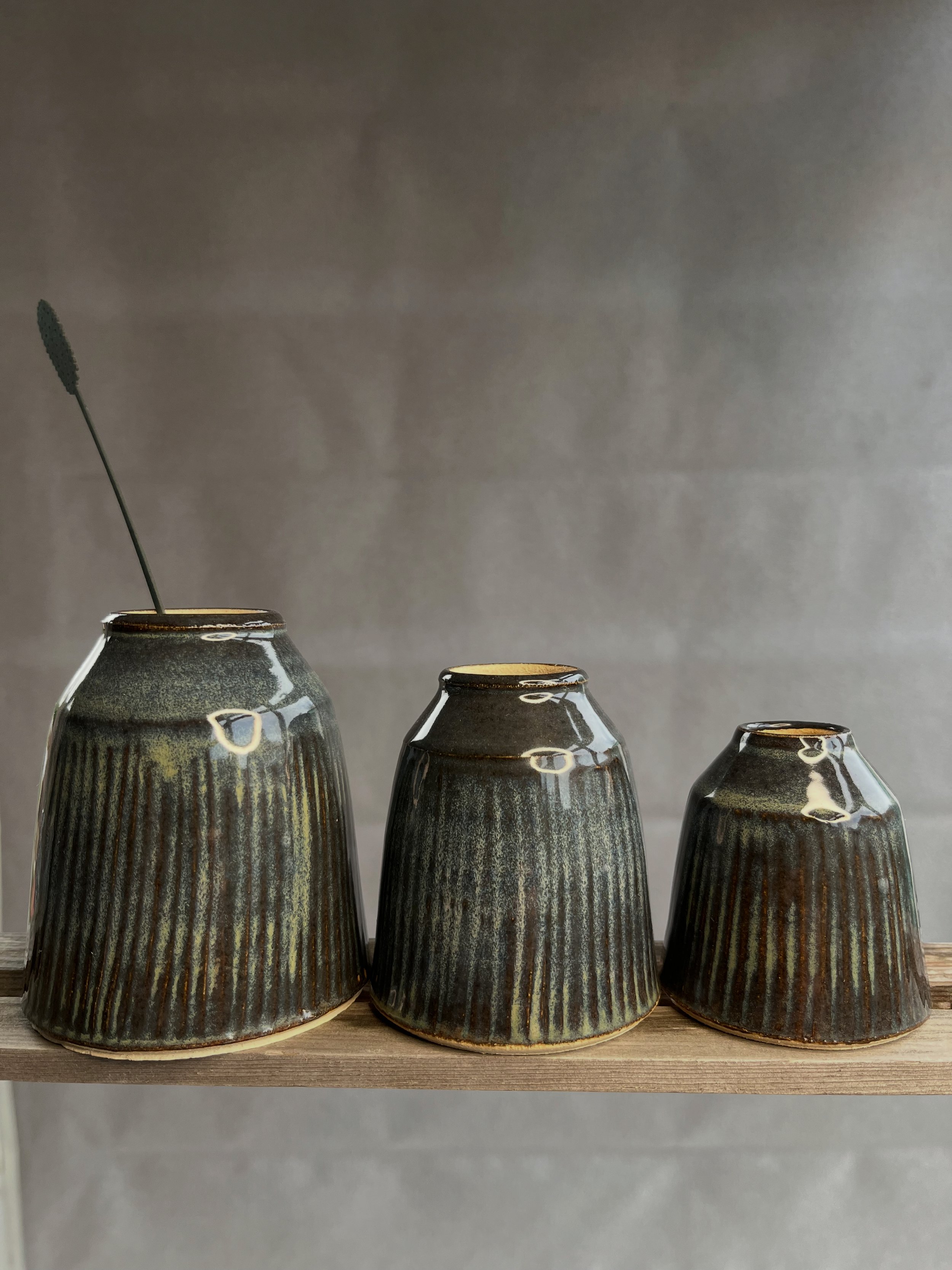 Three ceramic vases with a ribbed pattern and a dark, shimmery glaze, arranged on a wooden surface against a plain gray background.