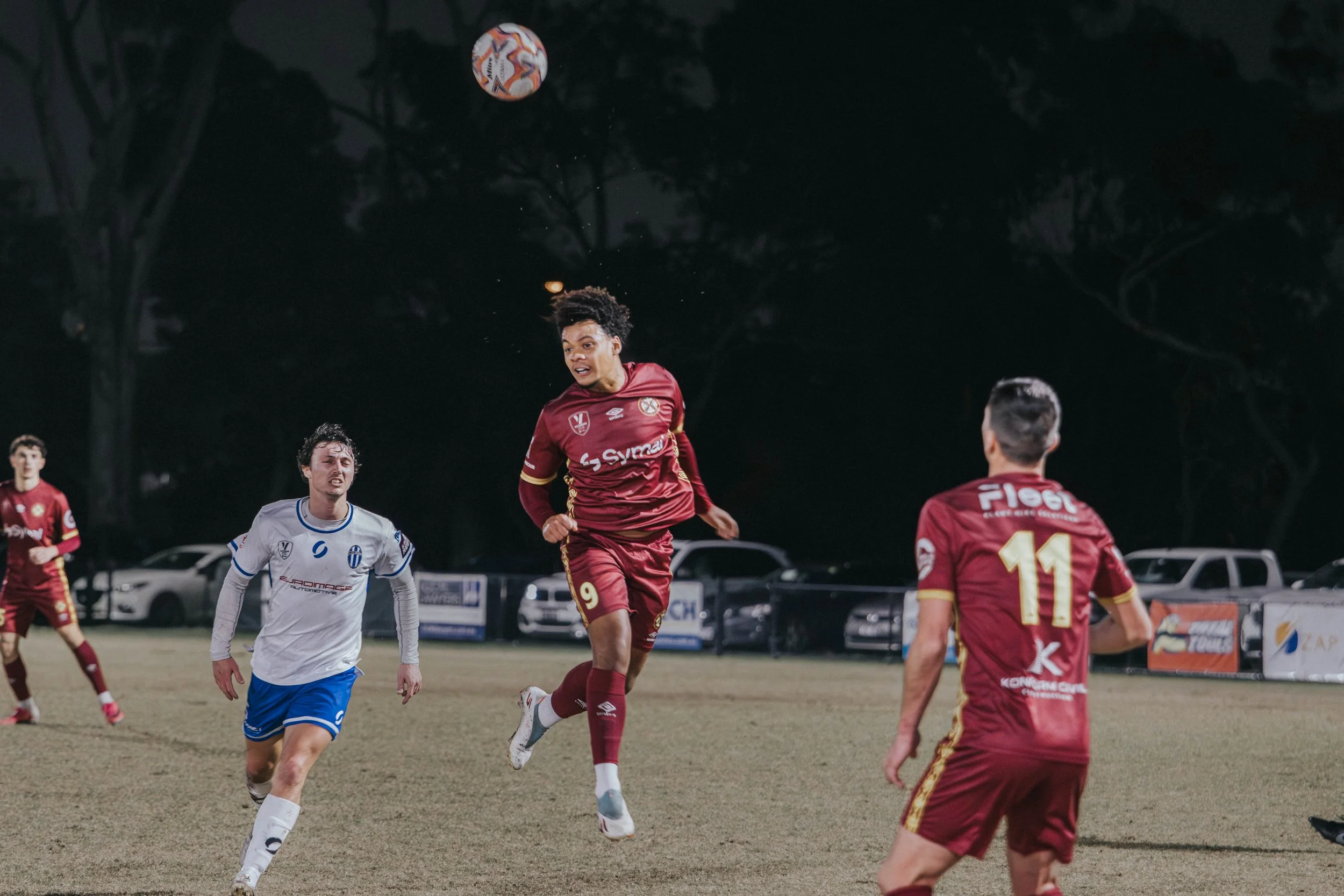 Soccer players during a match at night, with one player jumping to head the ball while others run and watch, on a field with parked cars and trees in the background.