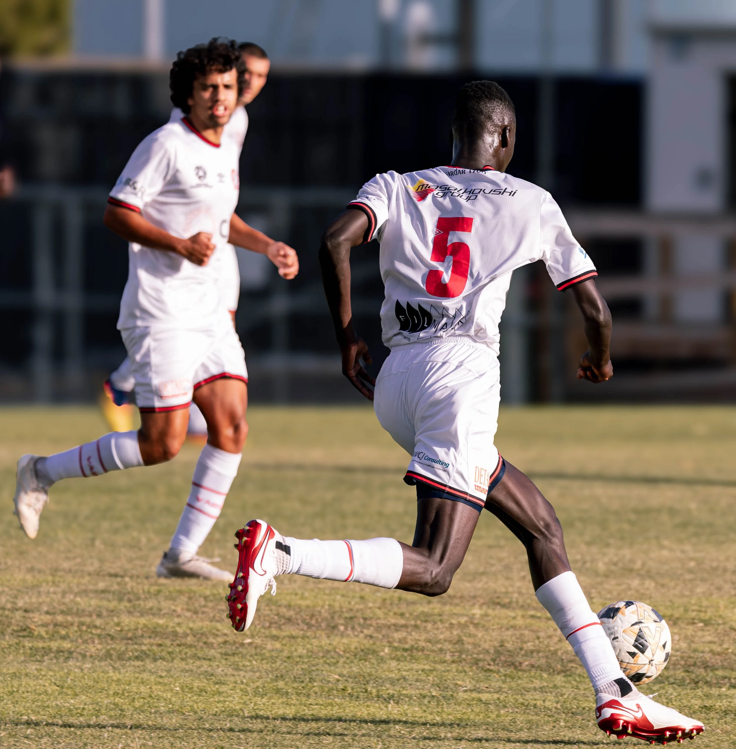 Three soccer players in white uniforms are on the field; one is lunging with the soccer ball at his feet, while the others are running behind him.