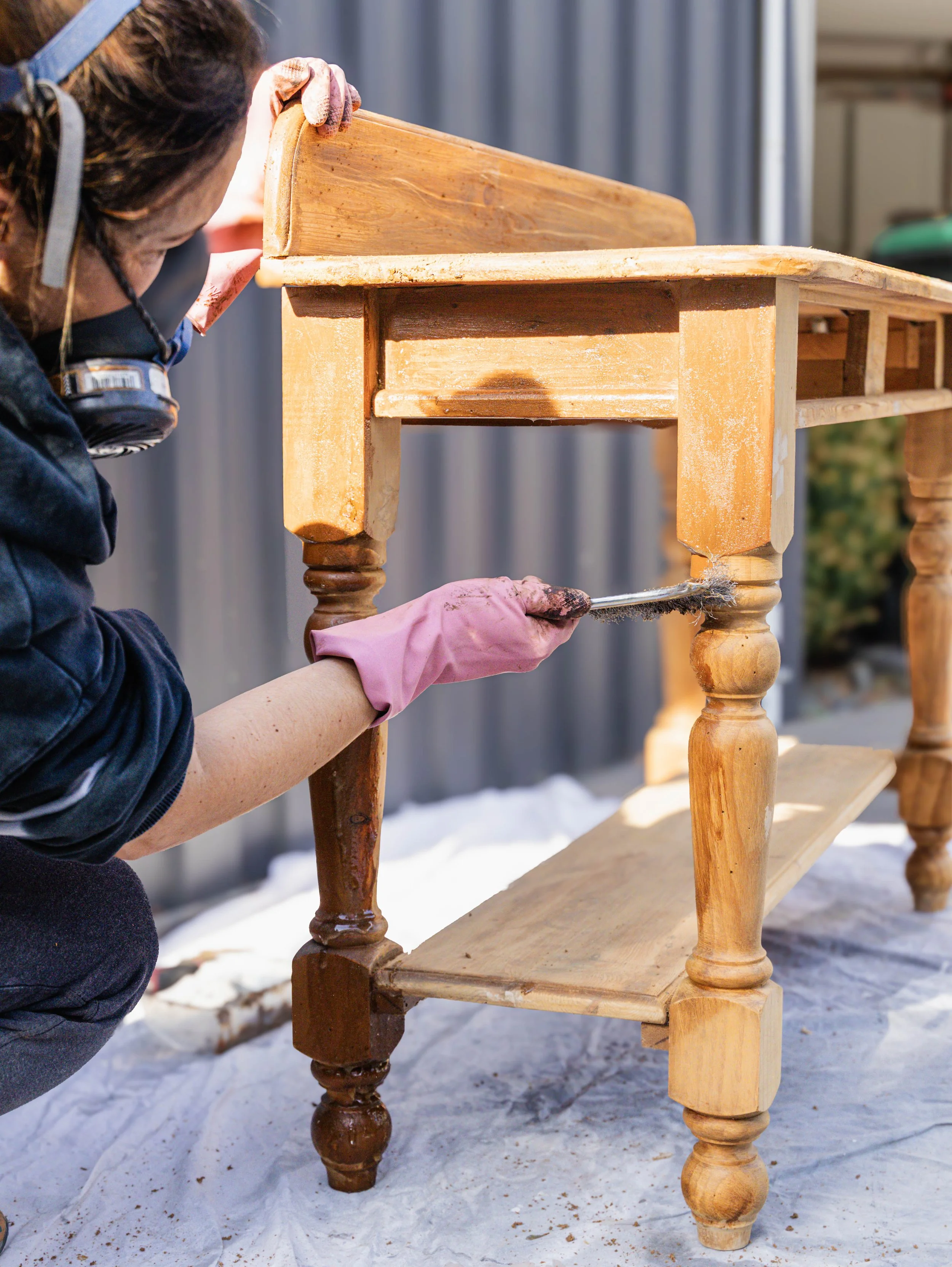 A person wearing gloves and a mask is sanding or restoring an old wooden chair outdoors, with snow on the ground.