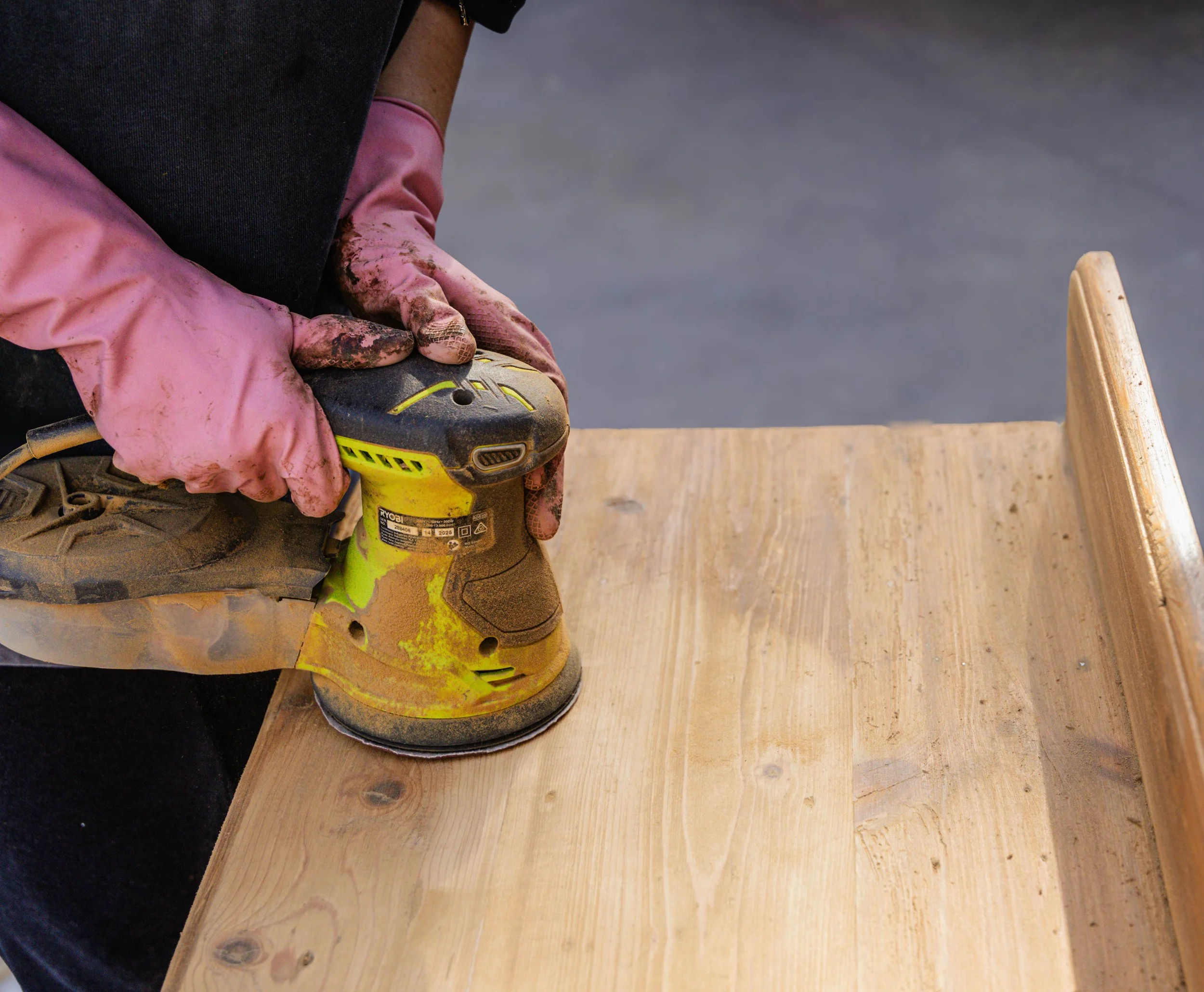A person in black clothing and pink muddy gloves using a yellow power sander on a wooden surface.