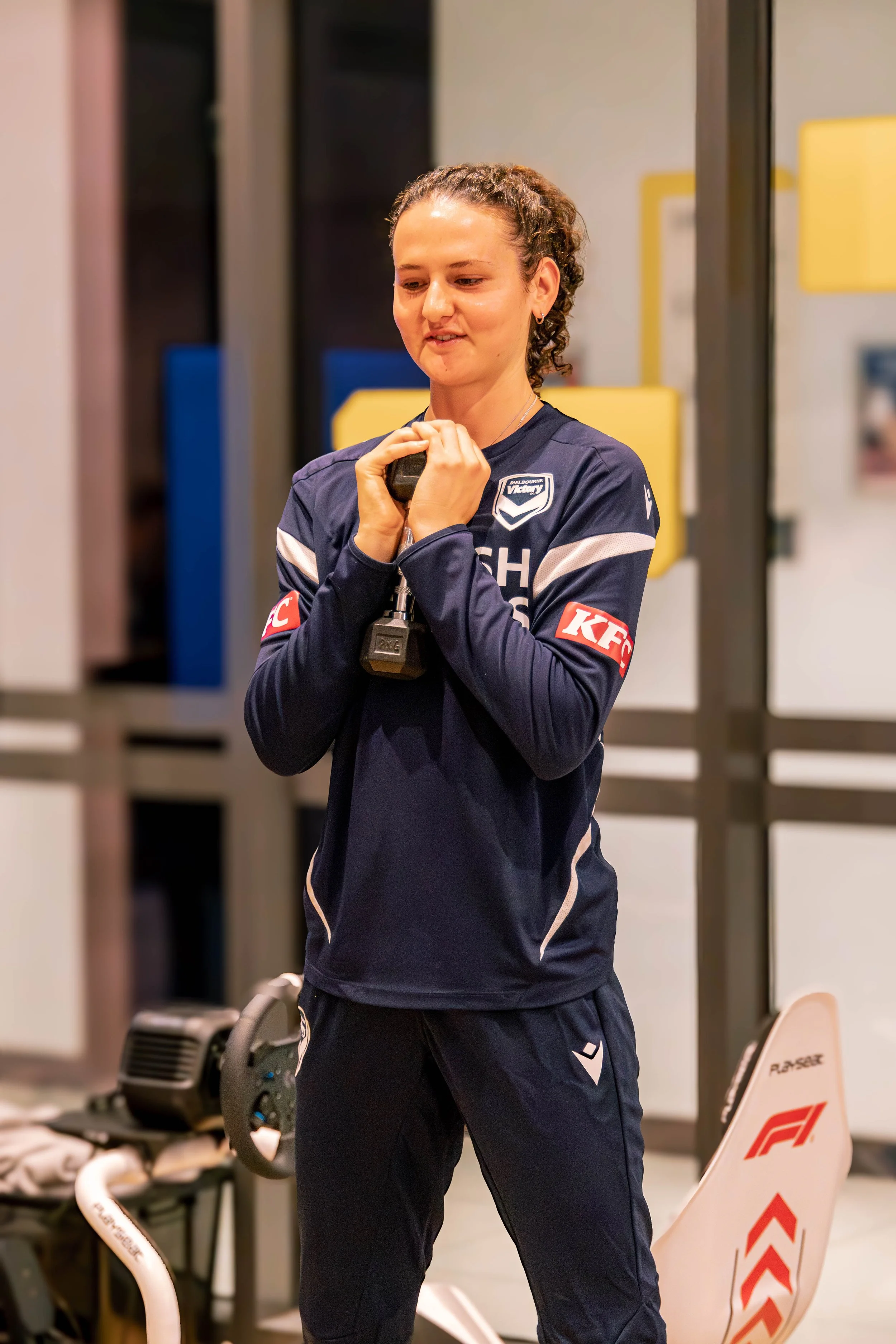 A woman in a dark blue sports uniform holding a dumbbell in a gym. The uniform has sponsorship logos including KFC and McDonald's. She is smiling slightly and appears to be stretching or warming up.