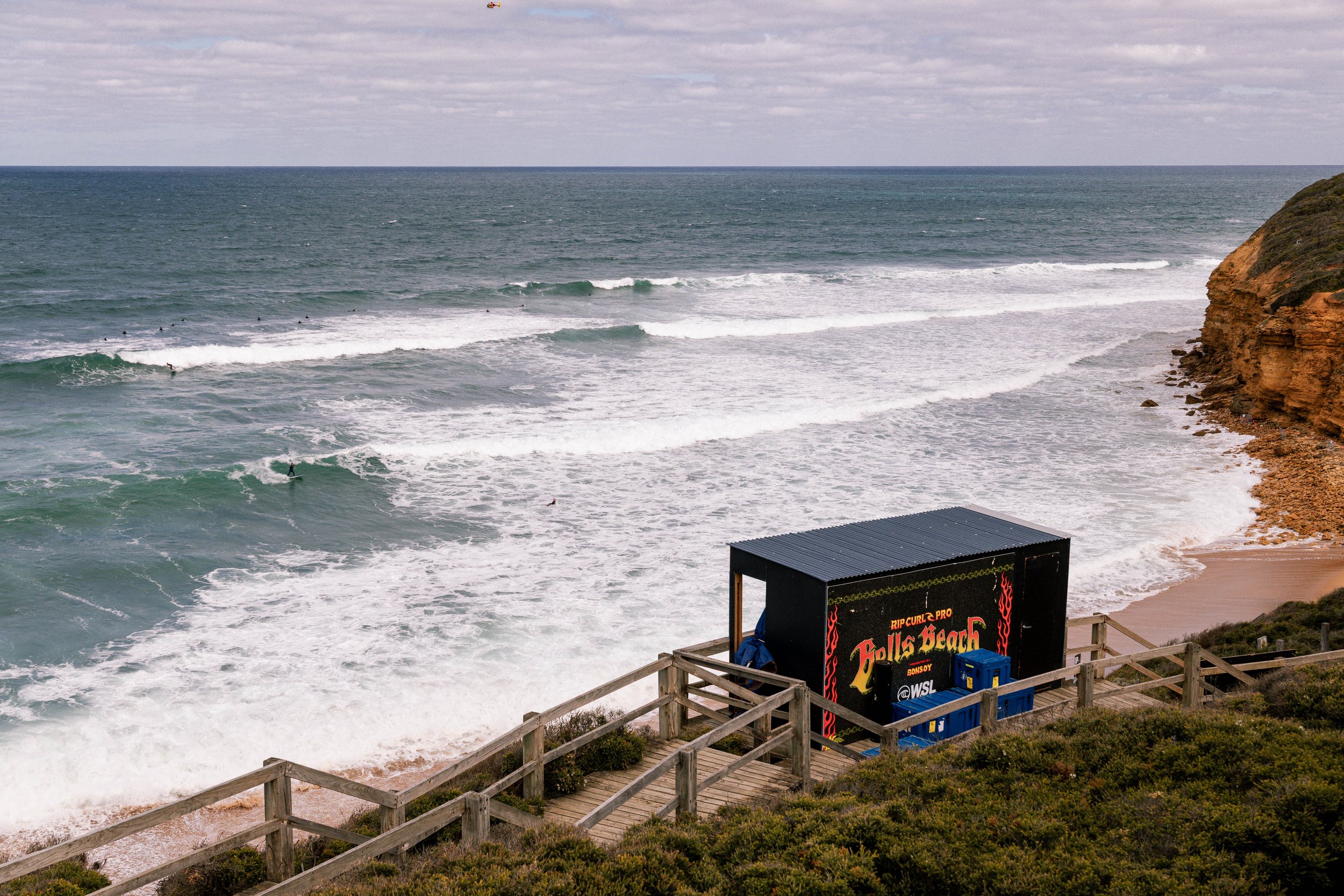 Ocean waves with surfers, a cloudy sky, a rocky cliff on the right, and a sign that reads 'Hollis Beach' near a wooden fence in the foreground.