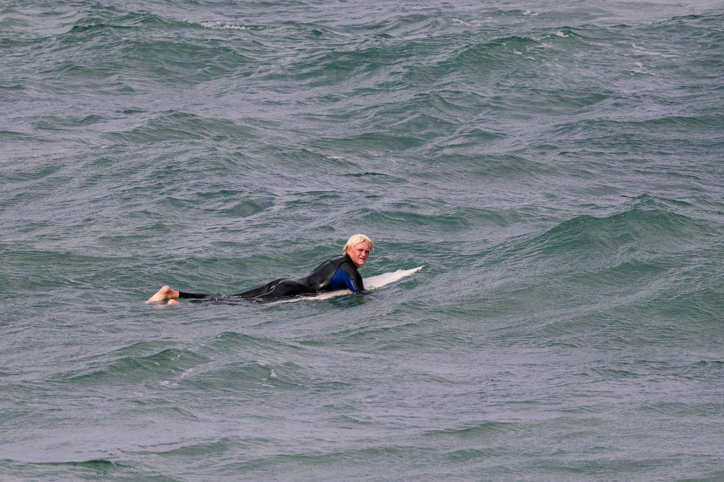 A person lying on a surfboard in the ocean, wearing a wetsuit and holding onto the surfboard, with waves around them.