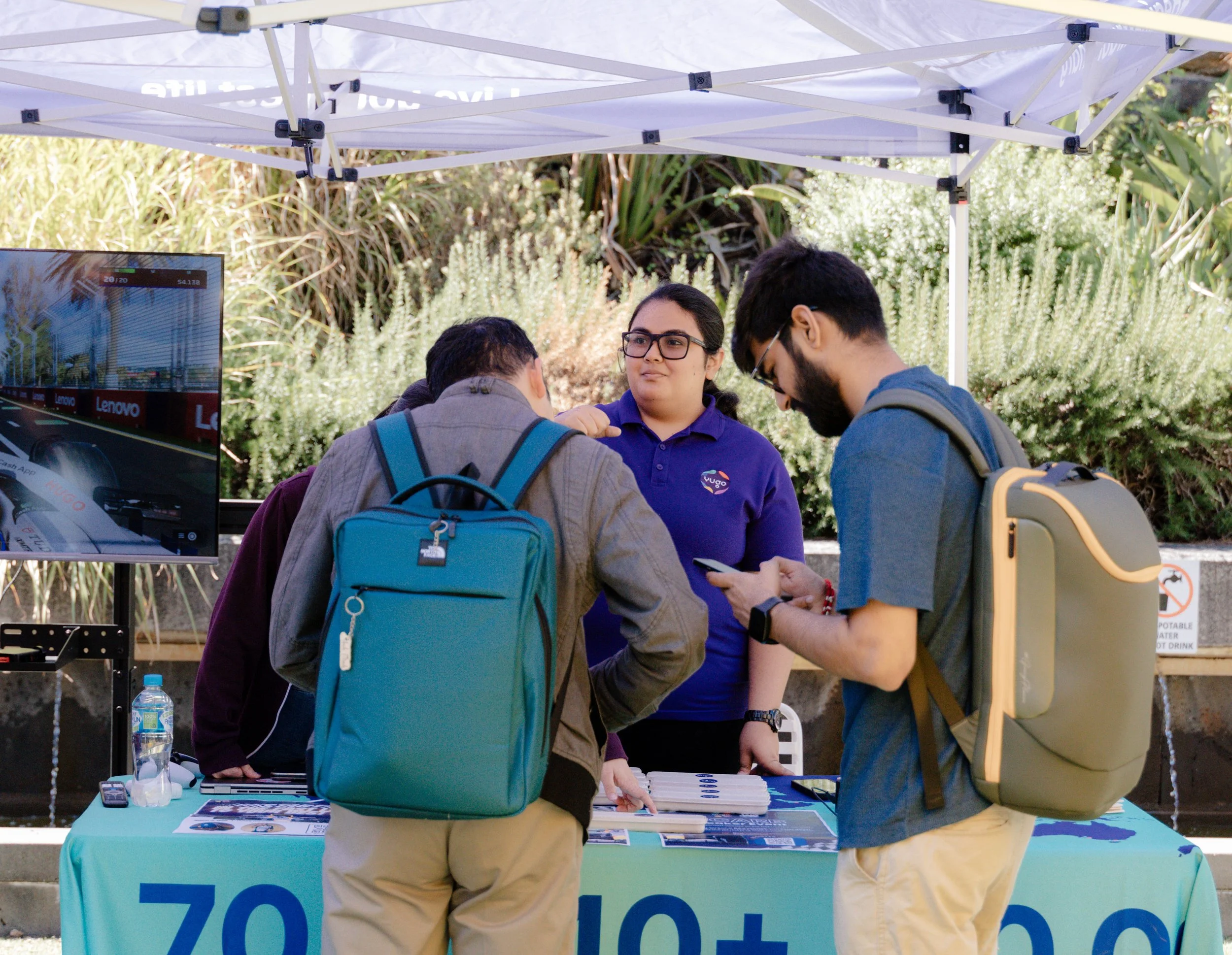 A group of people gathered around a booth with a person wearing a purple shirt and glasses engaging with others. There is a table with brochures and a water bottle, and a monitor displaying an image of a highway and the Lenovo logo. The booth is unde