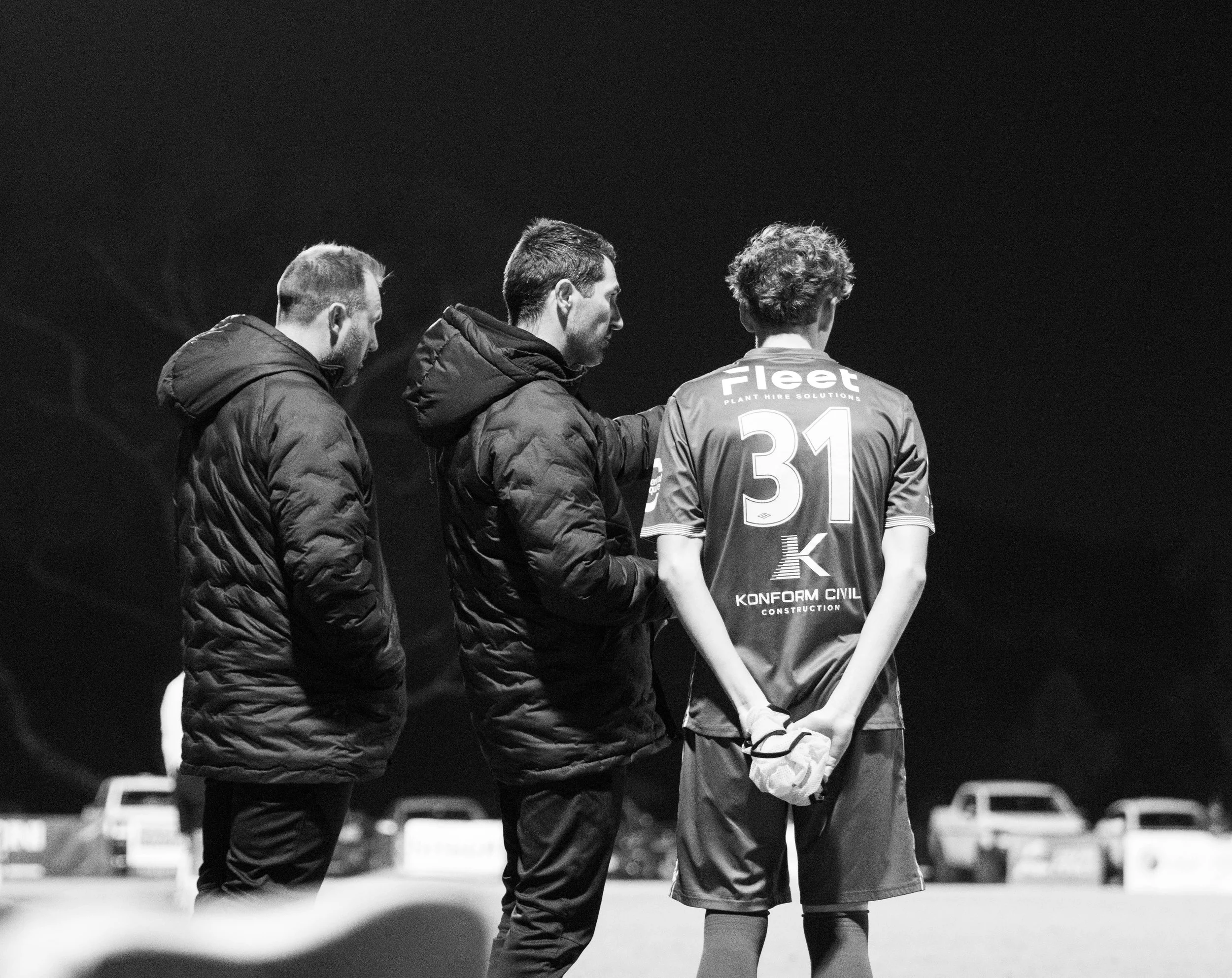 Three men, two in jackets and one in a soccer uniform with the number 31 and 'fleet' logo, standing on a sports field at night, viewed from behind.