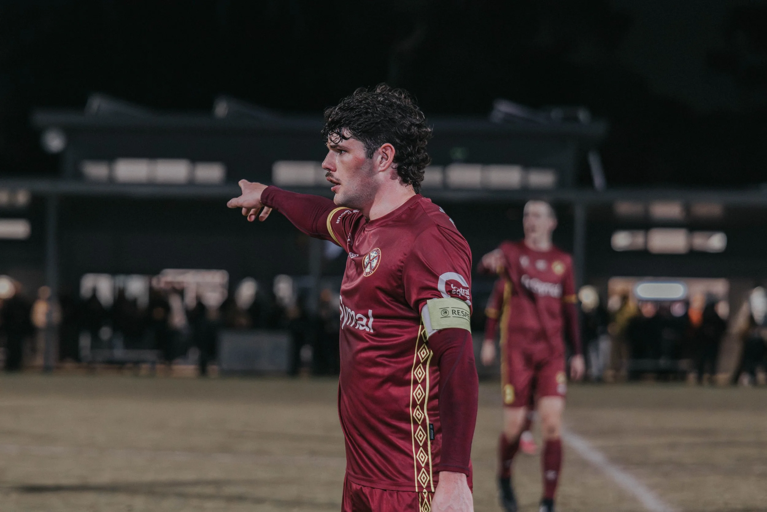 A male soccer player in a maroon jersey with gold trim and a captain's armband points with his right arm during a game. Other players in similar jerseys are visible in the background at a night match.