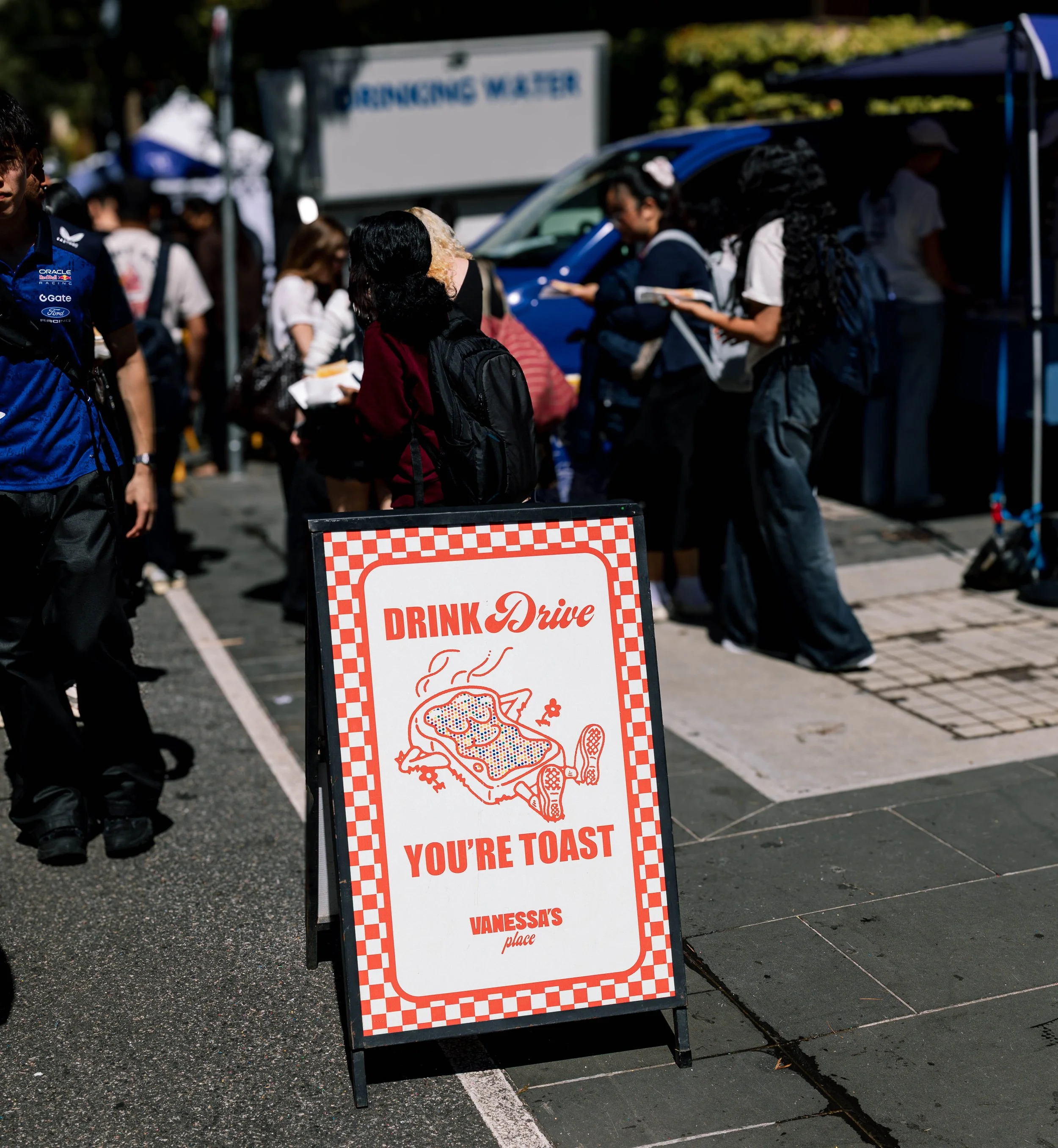 A sidewalk sign with a checkered red and white border advertising food at a drive-thru, with a cartoon image of a smiling steak and the text 'Eat Toast You're Toast Vanessa's Place'.