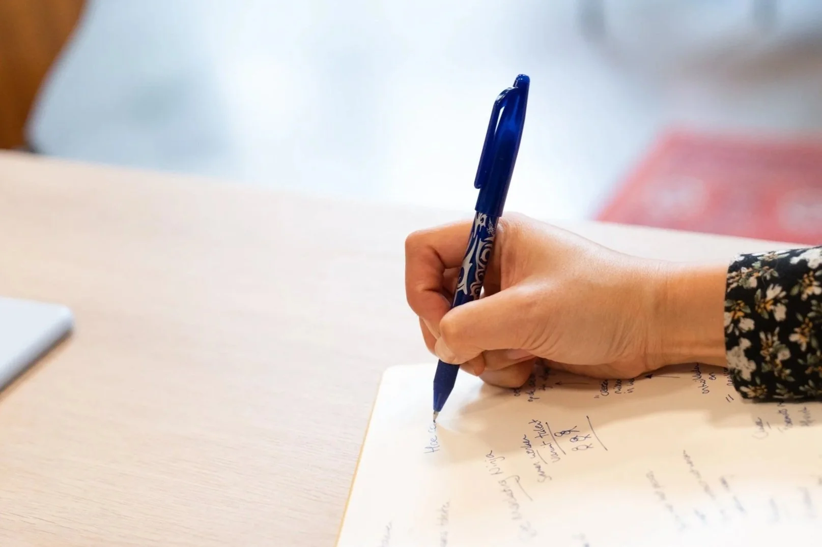 Een hand schrijft met een blauwe pen op een wit vel papier op een lichte houten tafel.