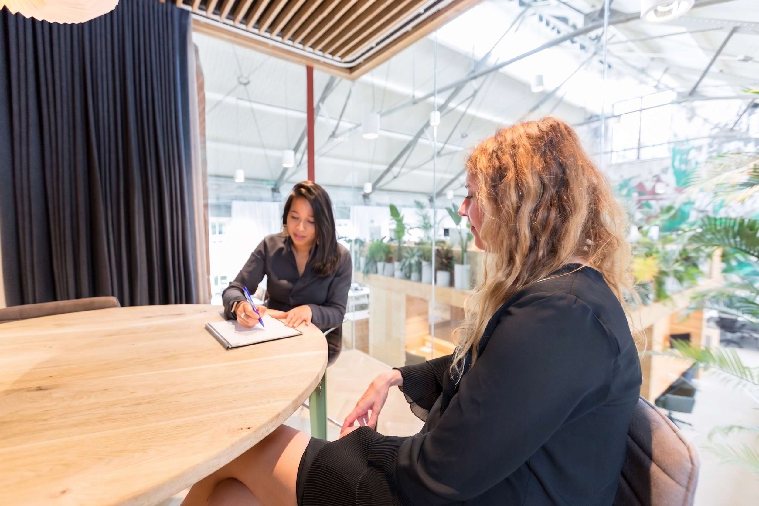 Twee vrouwen zitten aan een houten tafel in een moderne binnenruimte met veel planten op de achtergrond. De vrouw aan de linkerzijde schrijft in een boek terwijl de vrouw aan de rechterzijde naar haar kijkt.
