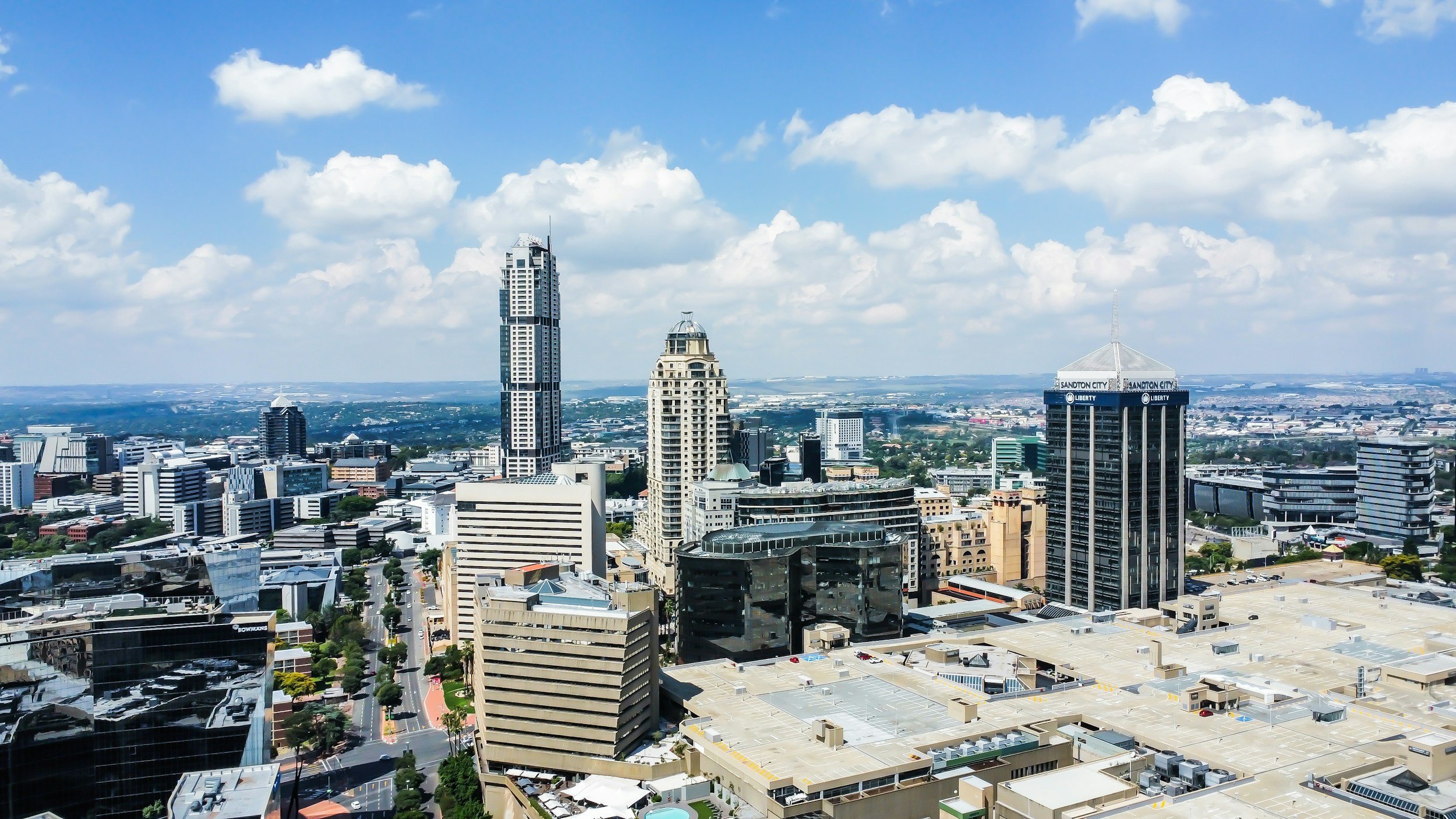 A city skyline with tall buildings, some labeled 'Sandton City' and 'Liberty', under a partly cloudy blue sky.