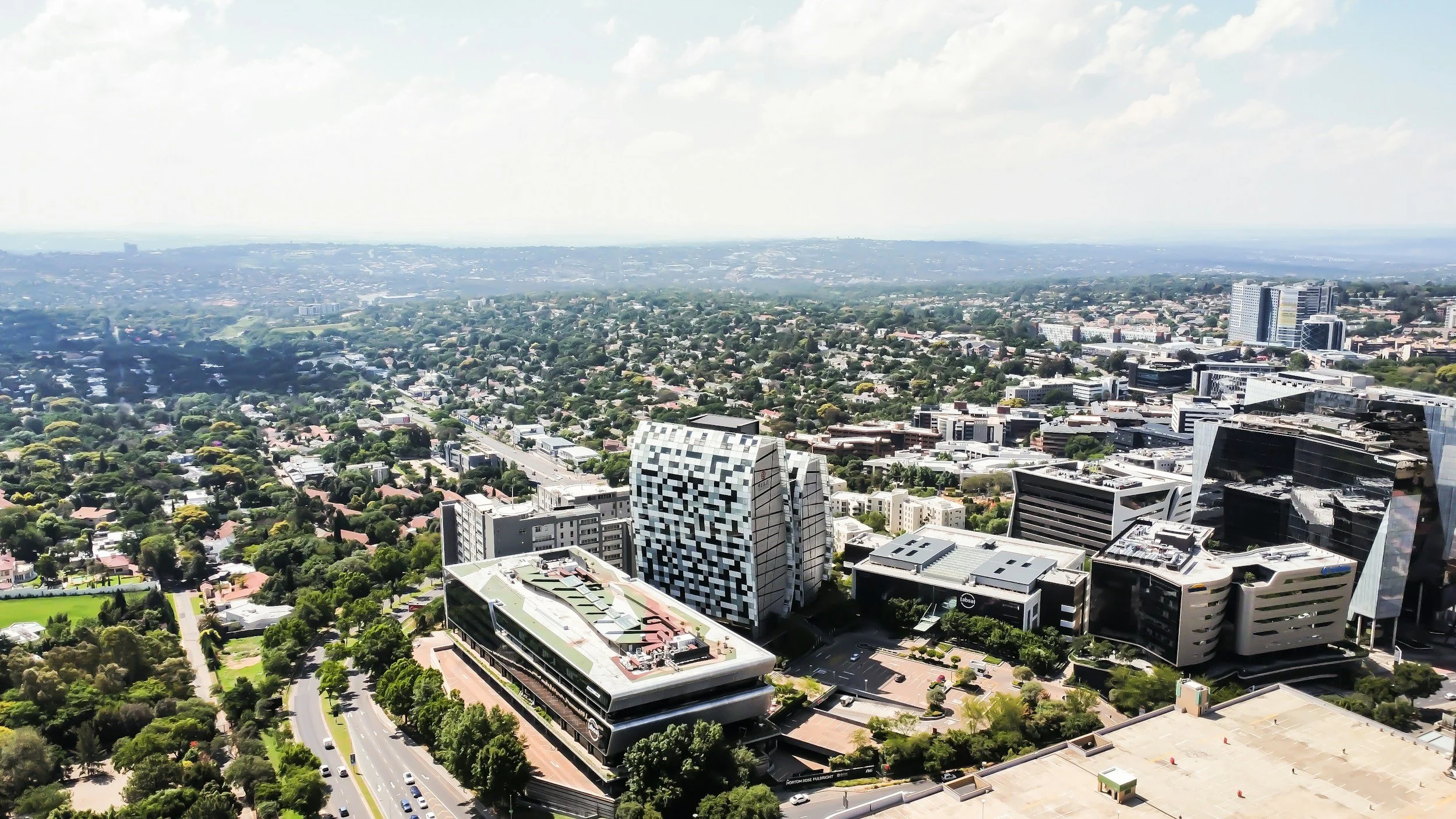 Aerial view of a city with tall modern buildings, roads, and green spaces extending into the distance under a partly cloudy sky.