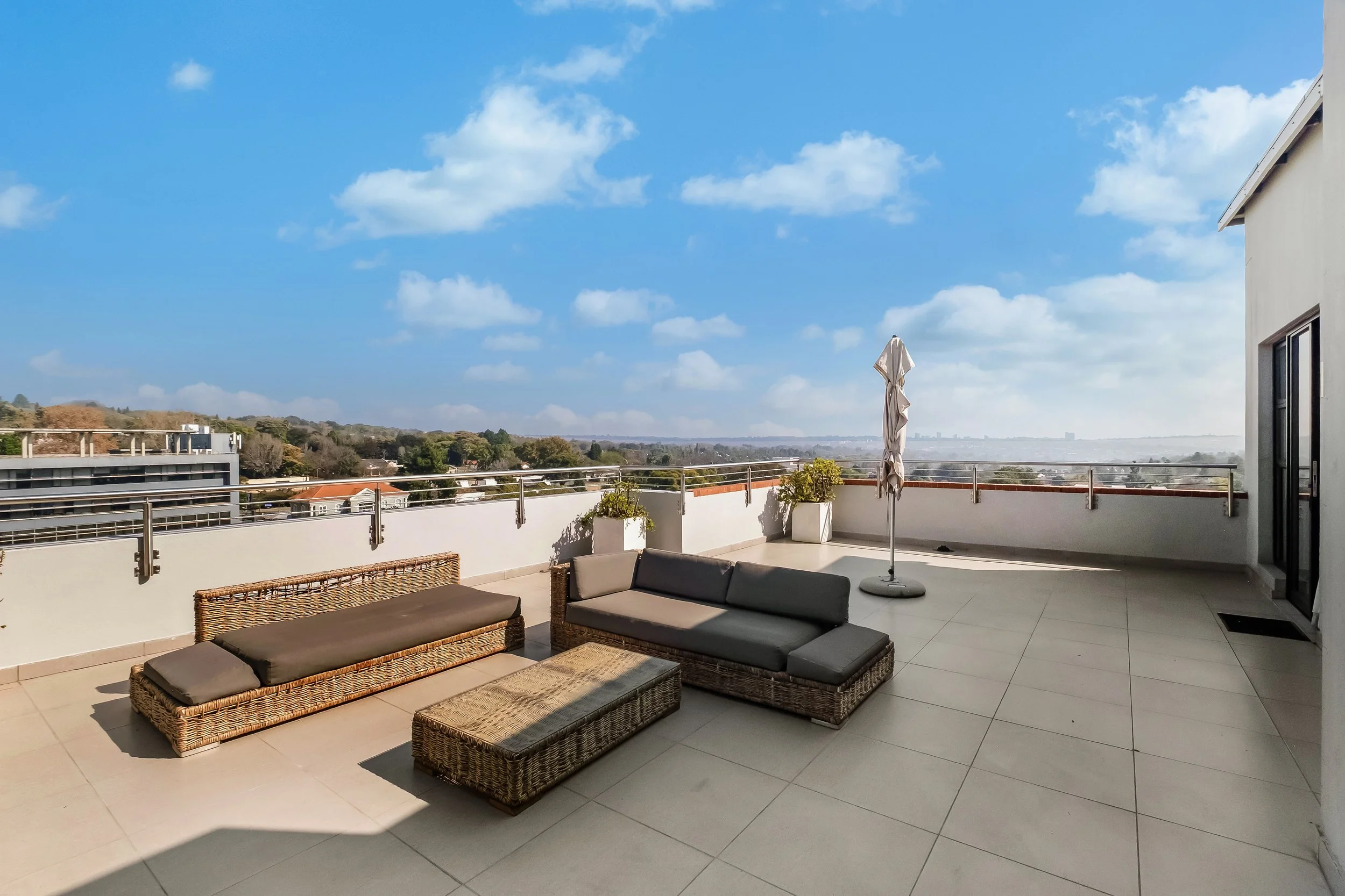 A spacious balcony or terrace with wicker outdoor furniture including a chaise lounge, a cushioned bench, and a small table. There is a closed patio umbrella and a few potted plants. The balcony has a metal railing and offers a panoramic view of a suburban landscape with trees, houses, and a partly cloudy blue sky.