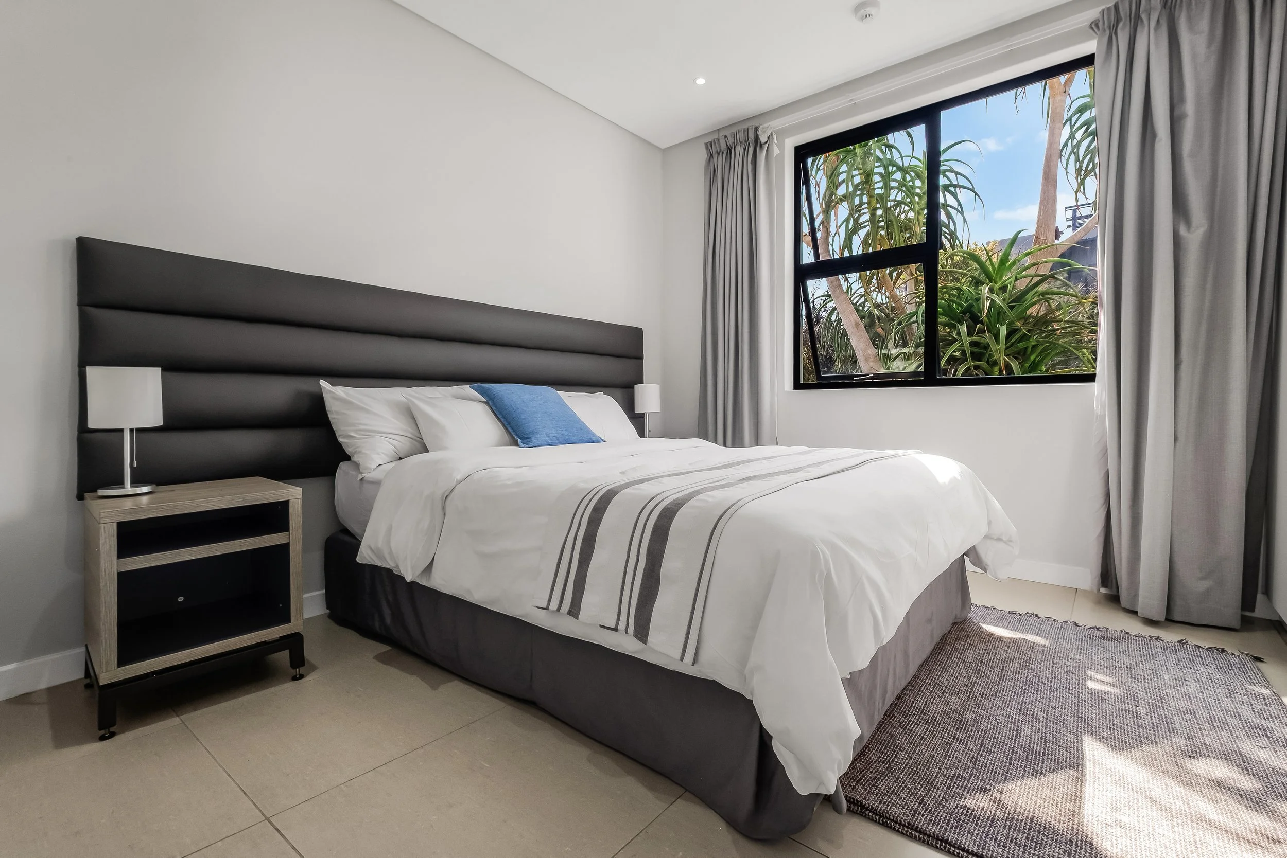 A bedroom with a bed, white bedding with gray stripes, and a blue pillow. There is a wooden nightstand with a white lamp on each side of the bed. A large window with gray curtains shows trees and blue sky outside.