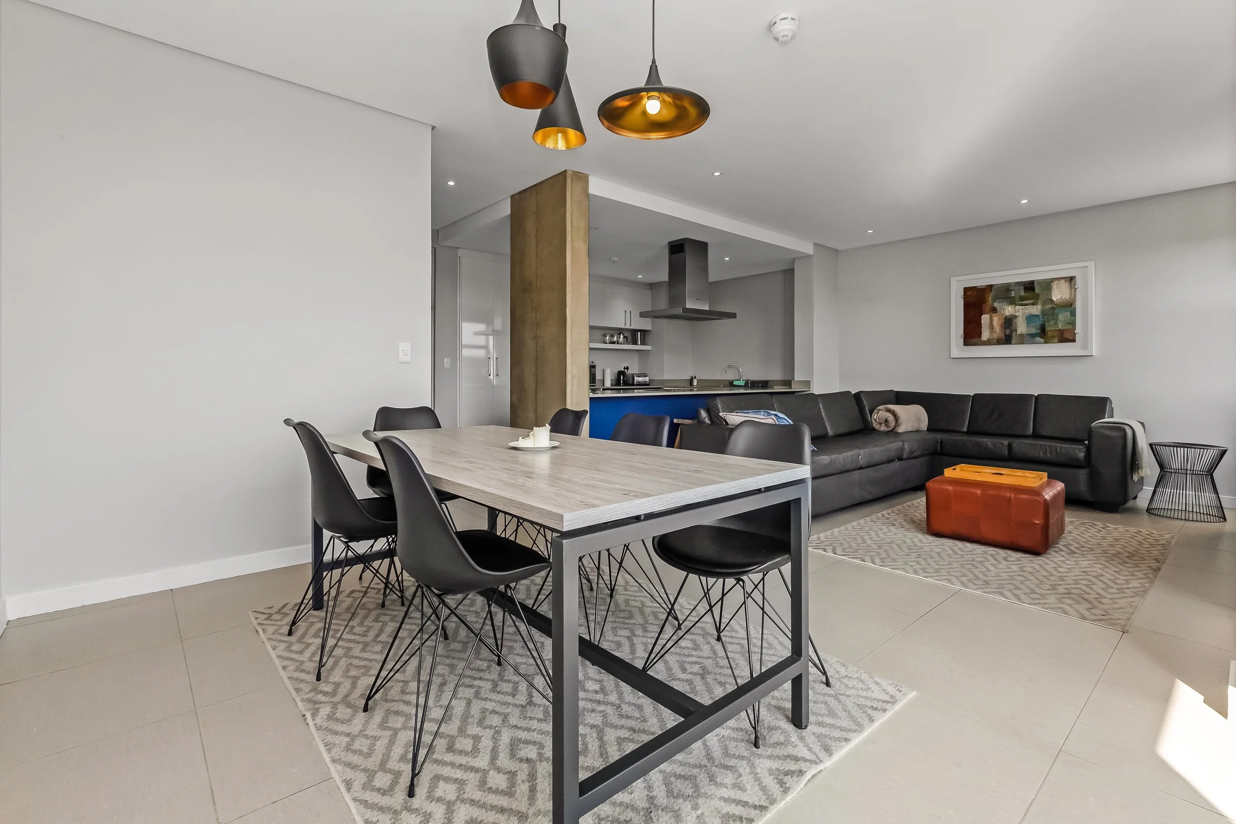 Modern open-concept living area with a dining table, black chairs, a black sectional sofa, a wooden and orange ottoman, and a kitchen with white cabinets, stainless steel appliances, and a blue accent on the kitchen island. There's a abstract painting on the wall and decorative pendant lights hanging from the ceiling.