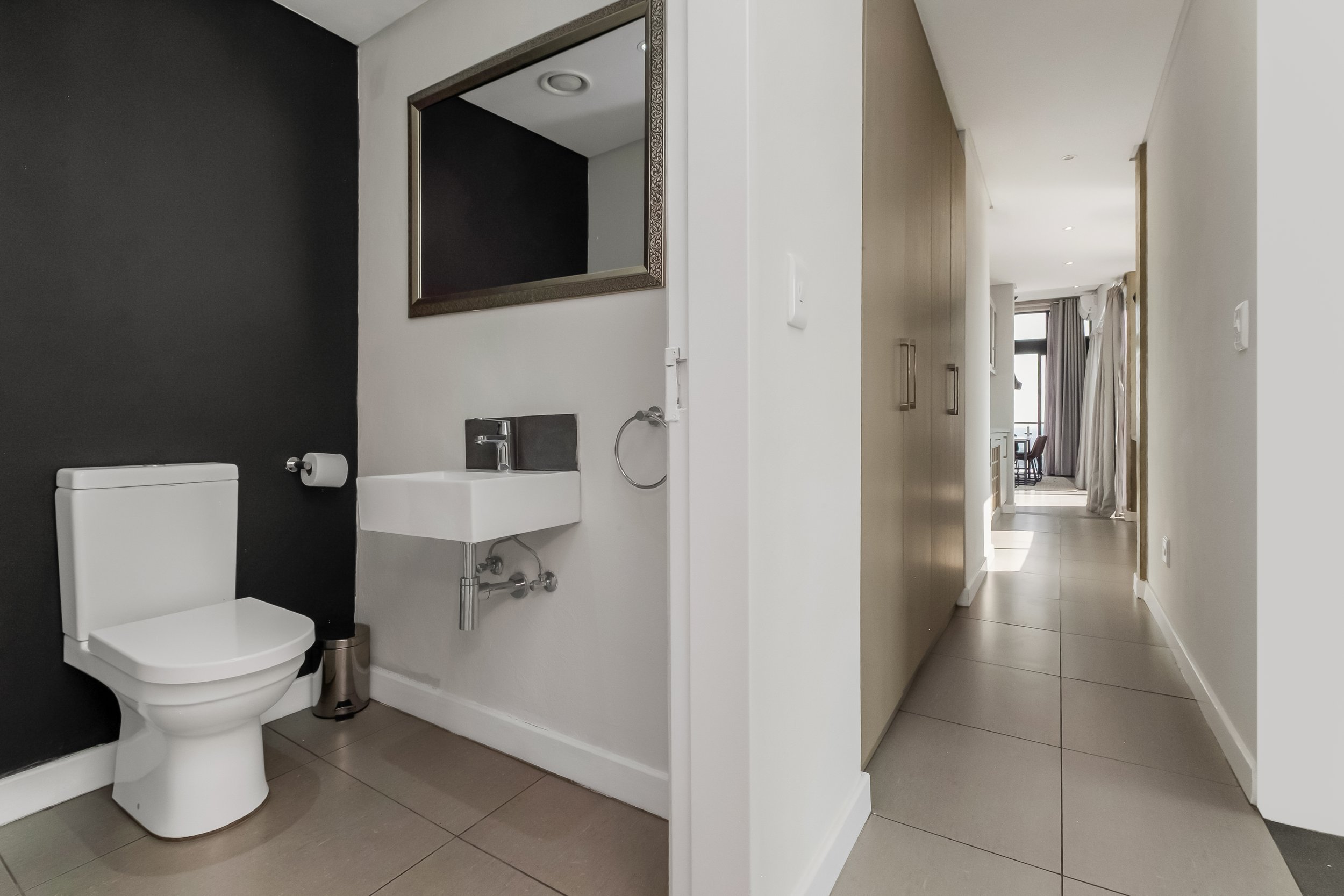 A modern bathroom with a black accent wall, a white toilet, a small white sink, and a mirror with an ornate frame. The bathroom leads into a hallway with wooden closet doors and a view into a living area with large windows and curtains.