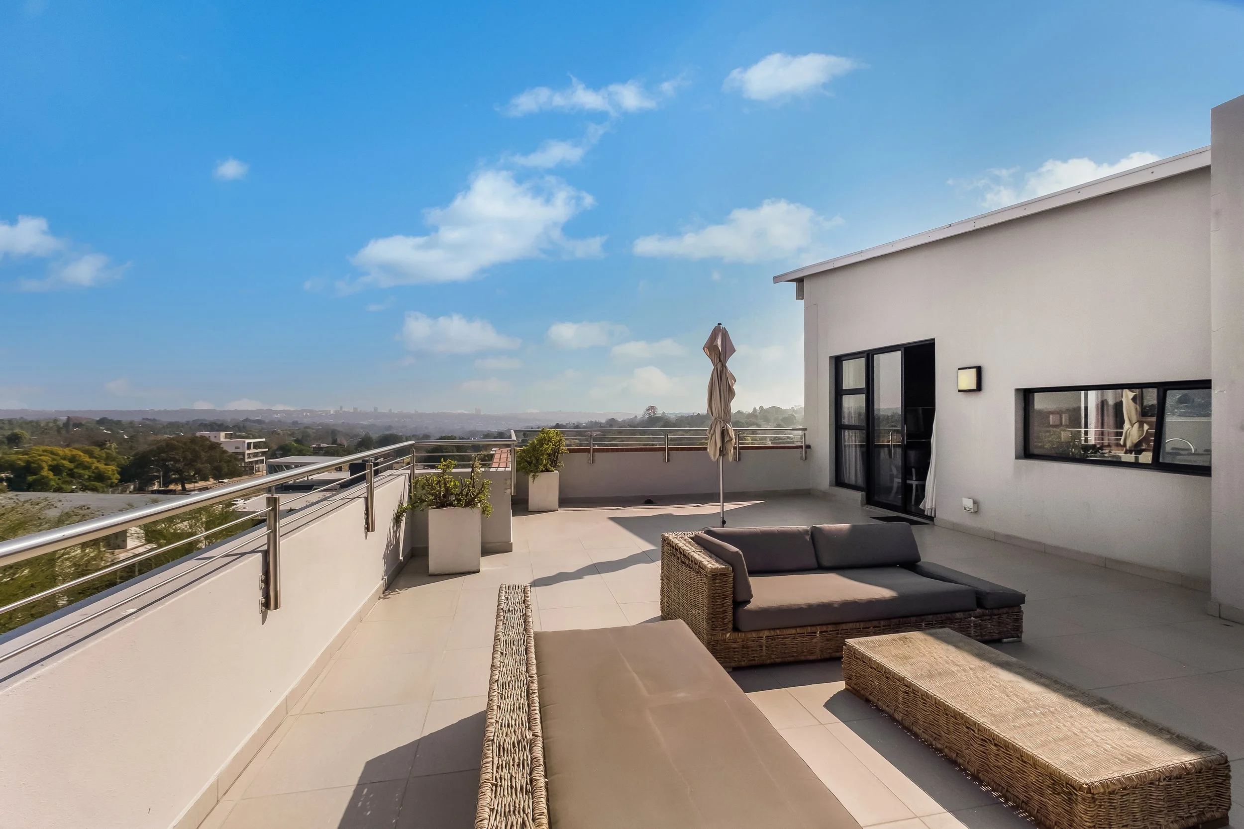 Rooftop terrace with outdoor furniture and potted plants, overlooking a city skyline and a partly cloudy blue sky.