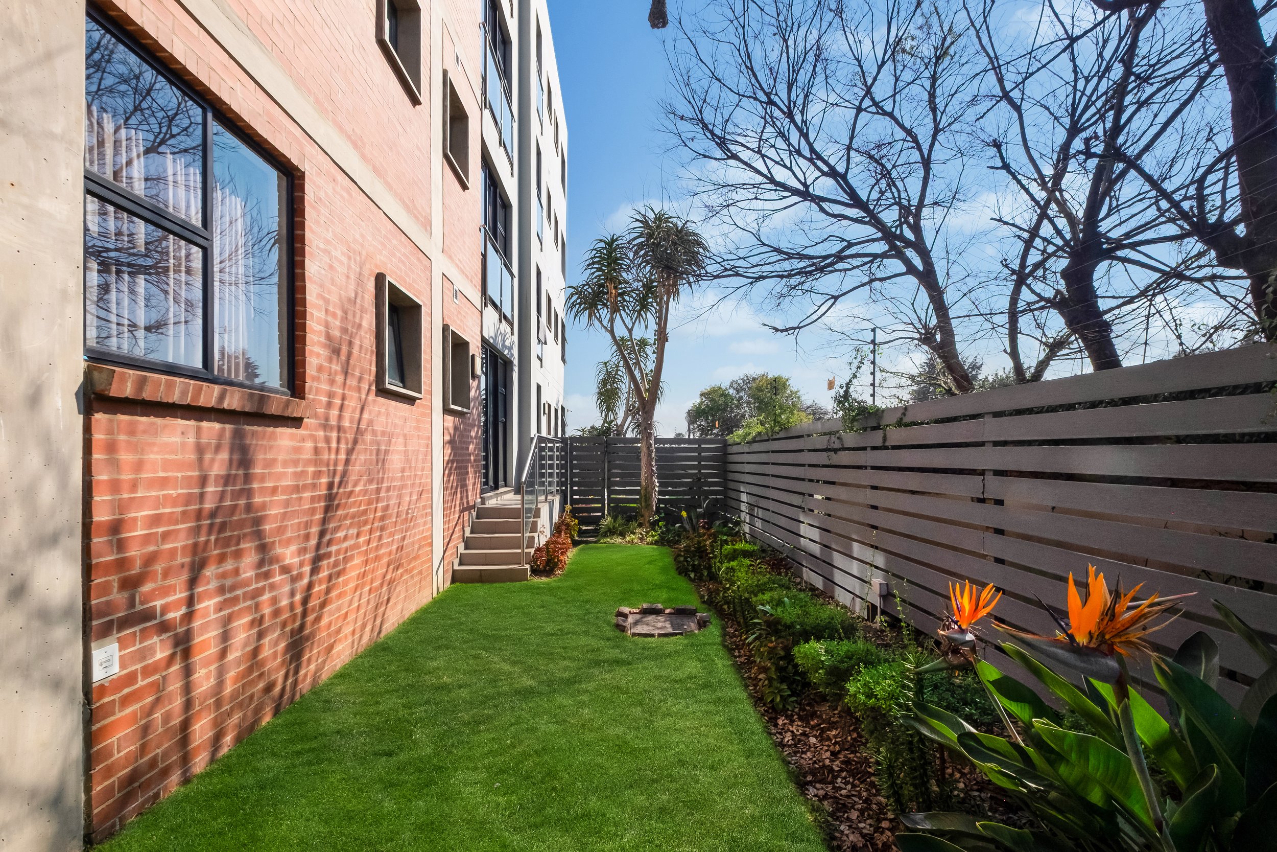 A small backyard with green artificial grass, with a red brick building on the left, a wooden fence on the right, and a clear blue sky with some branches of trees in the background.